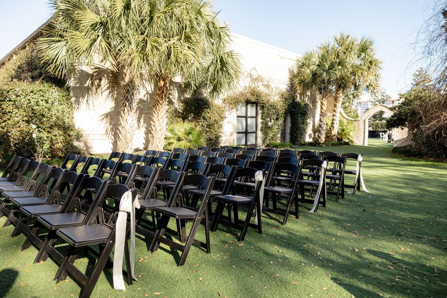 Rows of black folding chairs arranged on a lawn for an outdoor wedding ceremony with palm trees in the background.