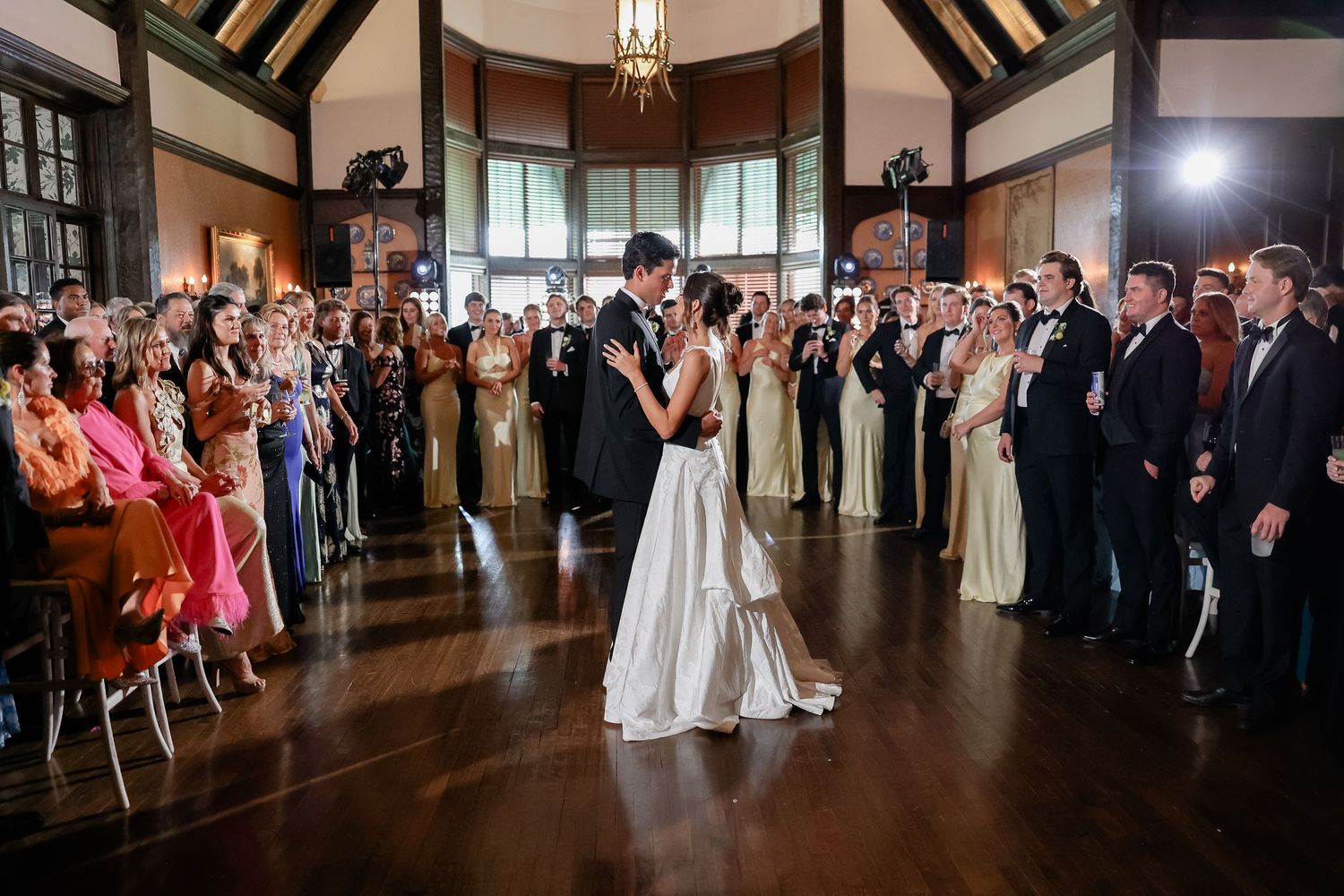 Bride and groom share first dance surrounded by wedding guests in elegant ballroom venue.