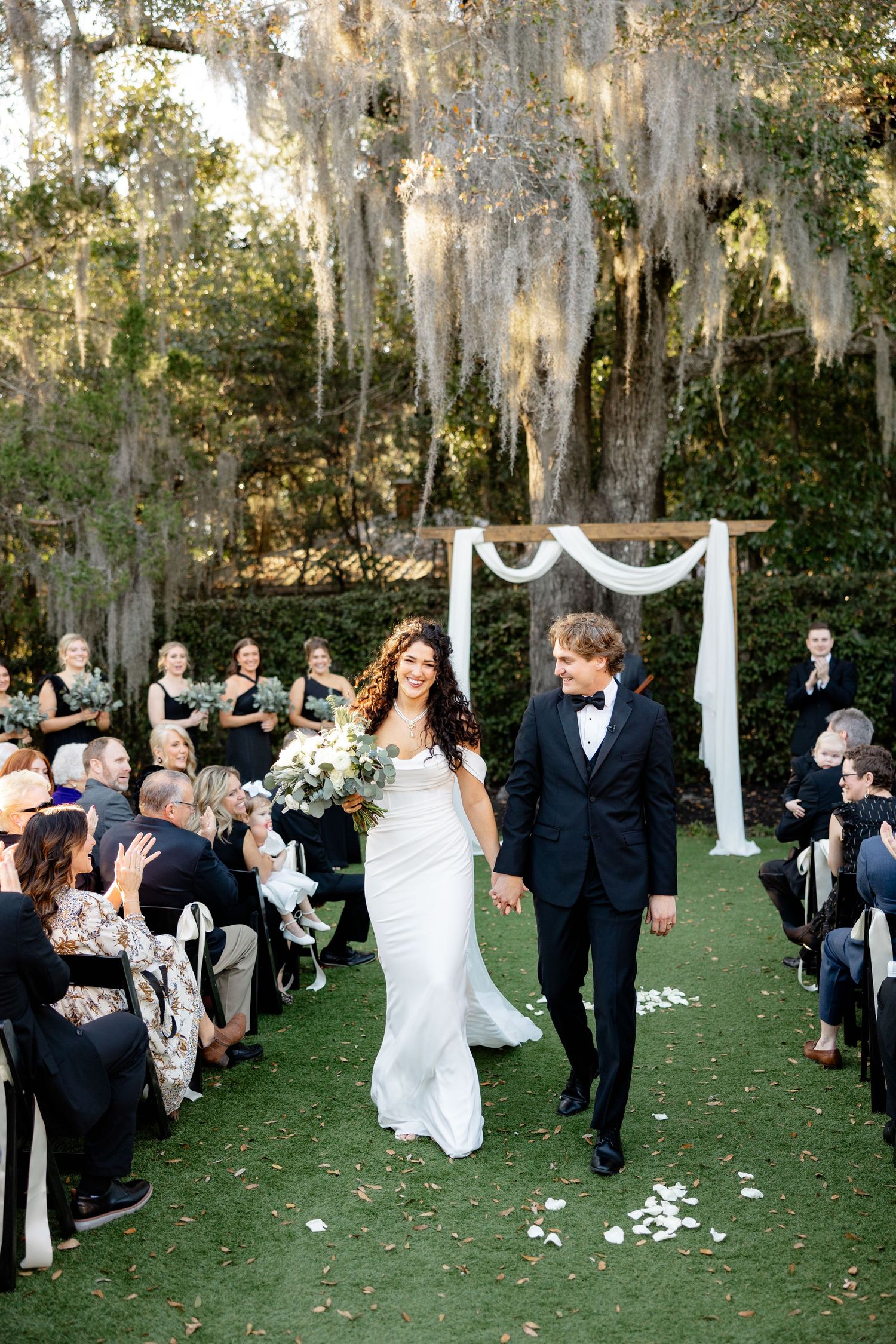 Bride and groom walk down outdoor aisle under Spanish moss-draped trees after wedding ceremony.