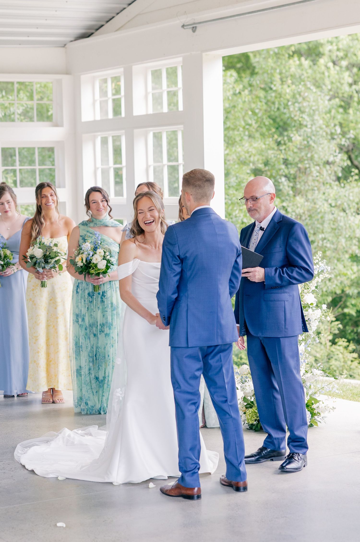 Bride laughing joyfully during outdoor wedding ceremony with bridesmaids in blue and yellow dresses beside her.