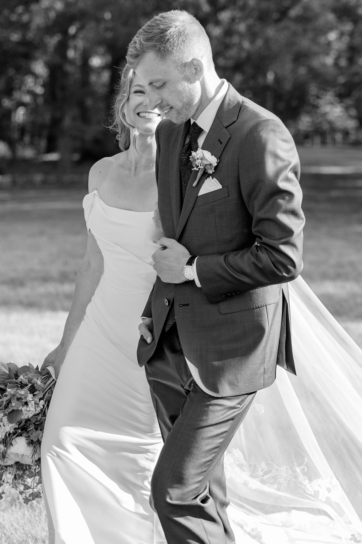 Black and white photo of a smiling bride in a flowing gown walking arm-in-arm with a groom in a suit outdoors.