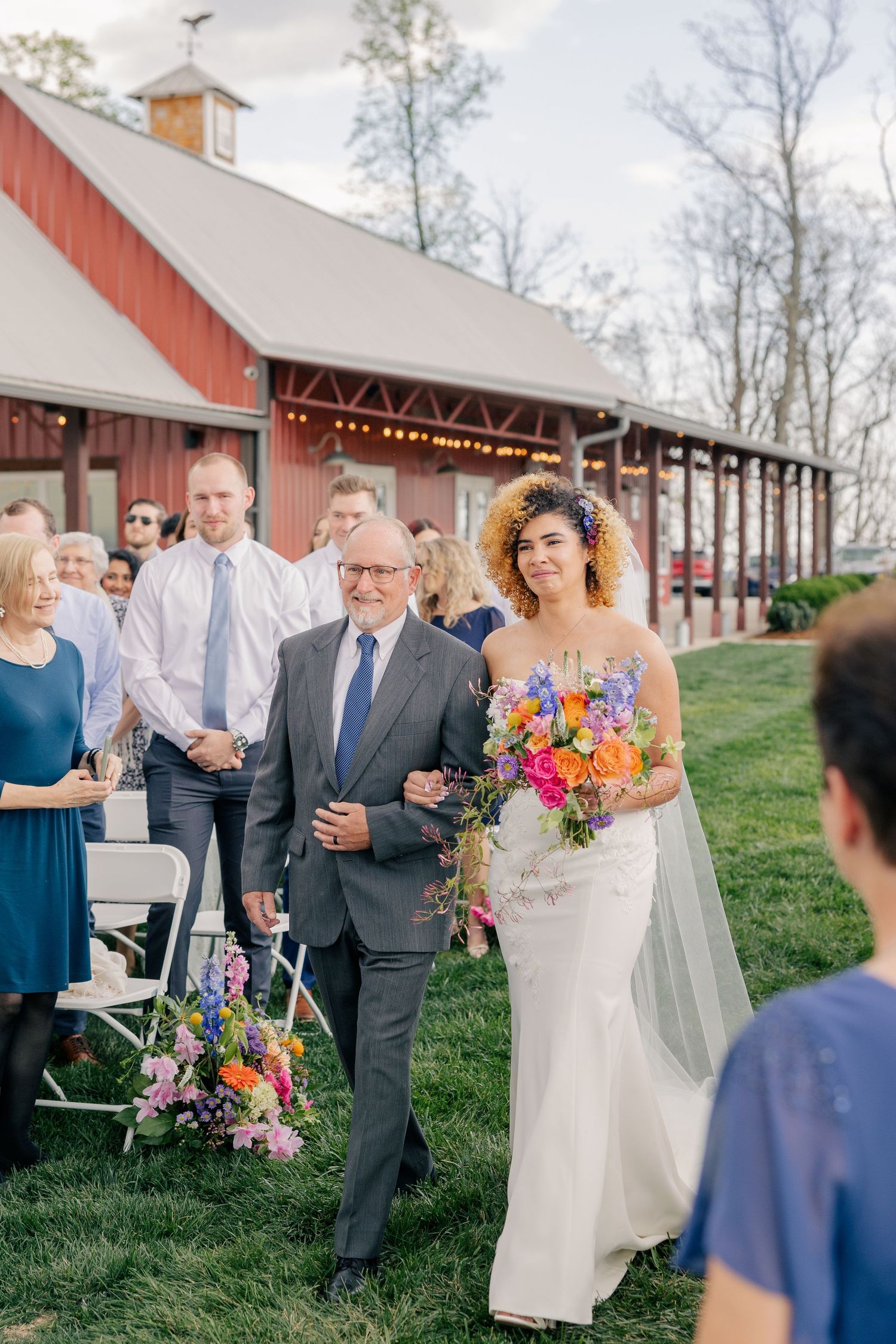 Bride in white gown walks down aisle with escort holding vibrant bouquet past guests near red barn venue.