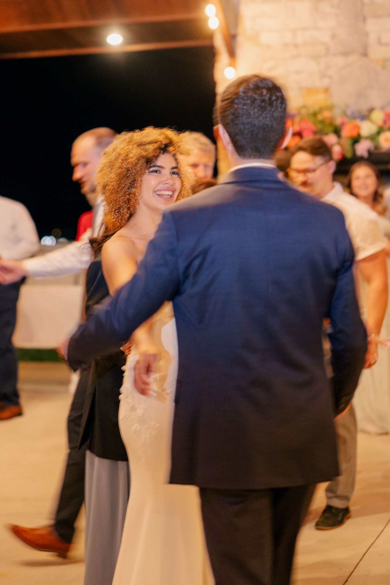 Smiling bride dances with groom in blue suit during evening wedding reception under warm string lights.