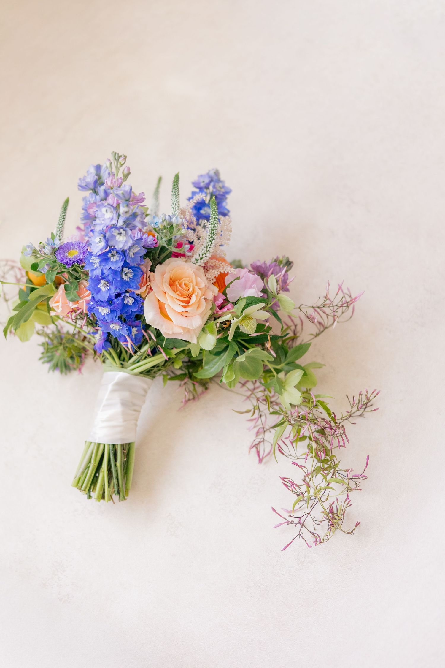 Bridal bouquet with blue delphinium, peach roses, and wildflowers on white background.