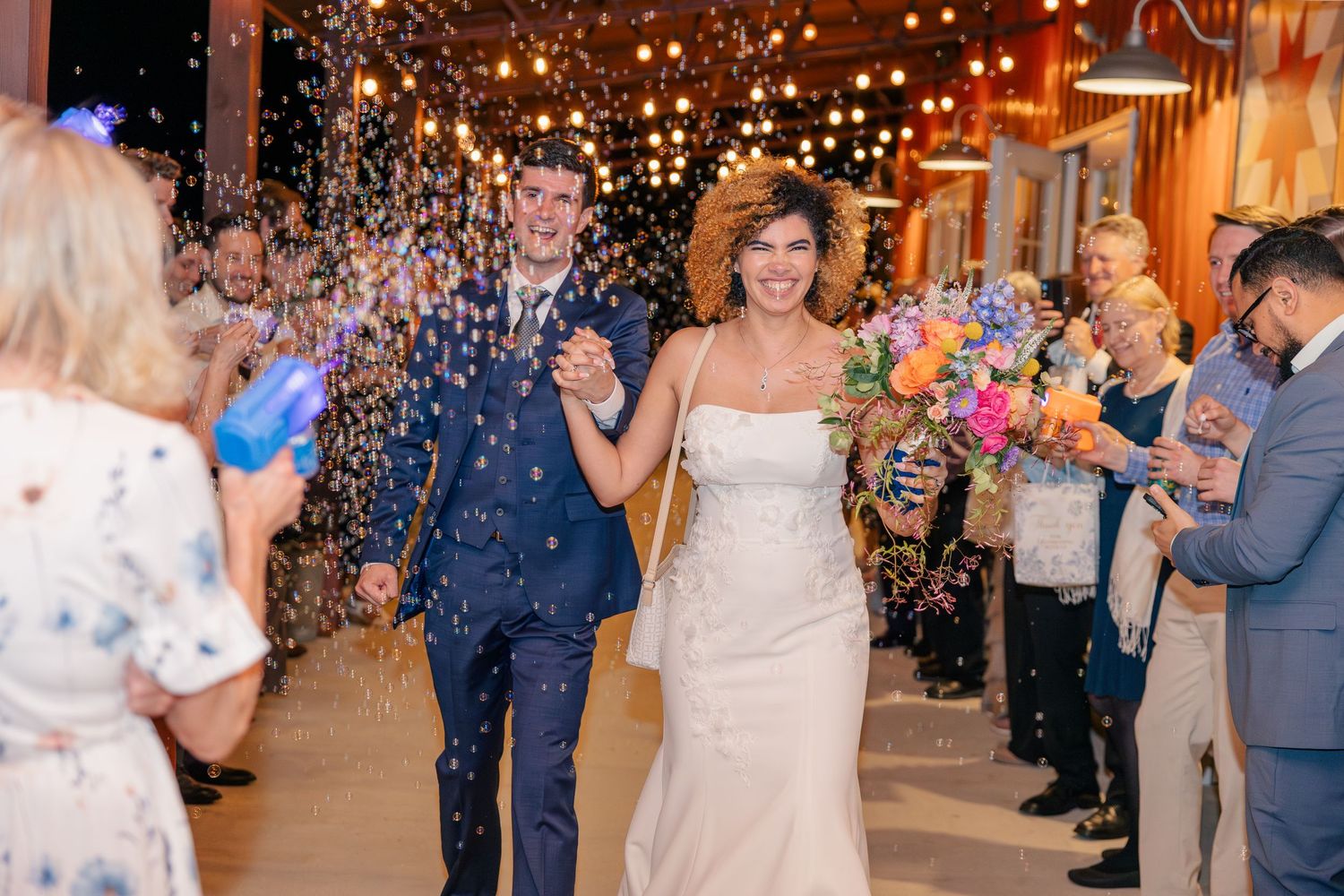 Newlyweds walk through confetti celebration with guests cheering inside a rustic wedding reception venue.