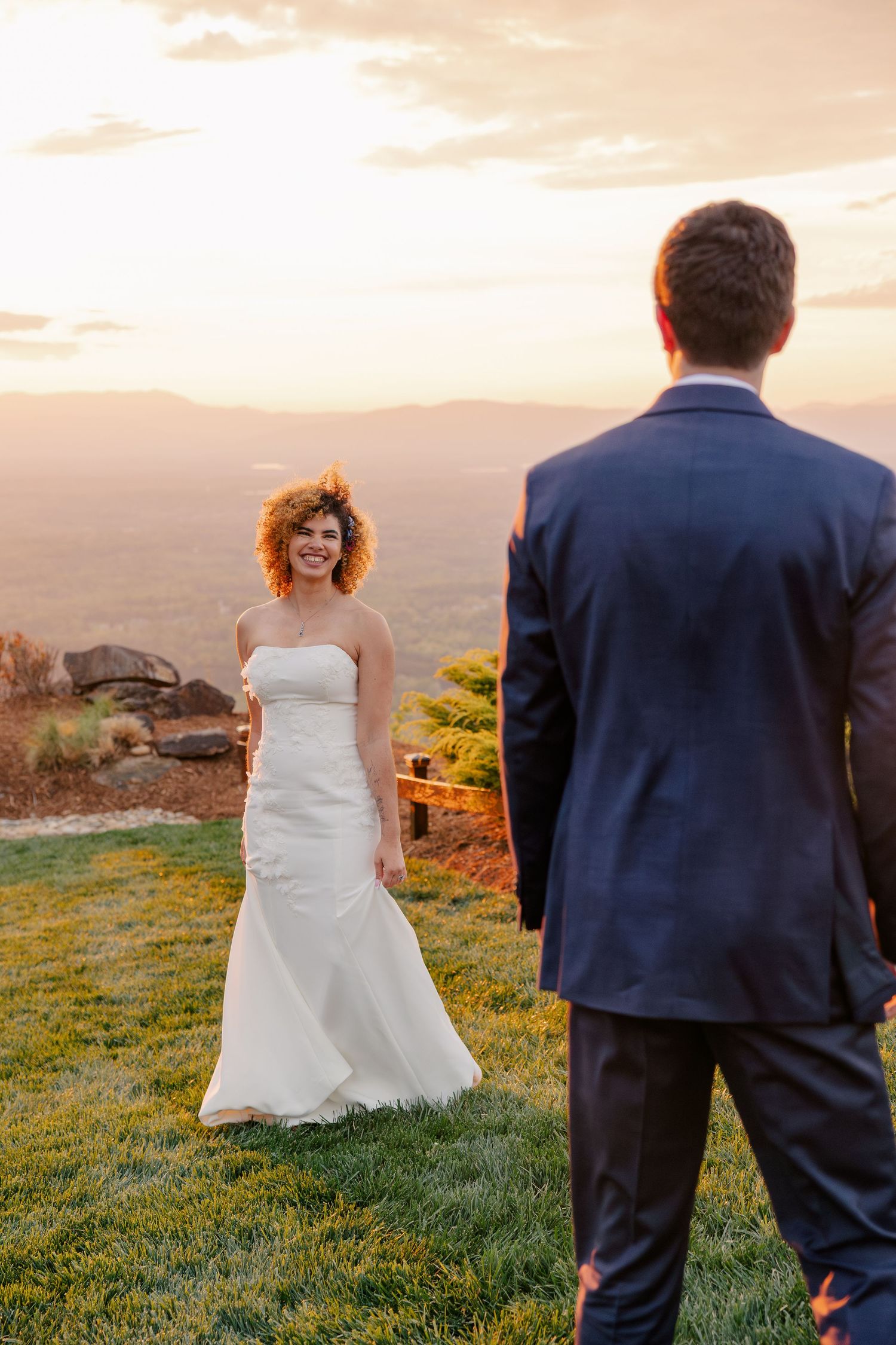 Smiling bride in white strapless gown walks toward groom at golden hour on scenic overlook.