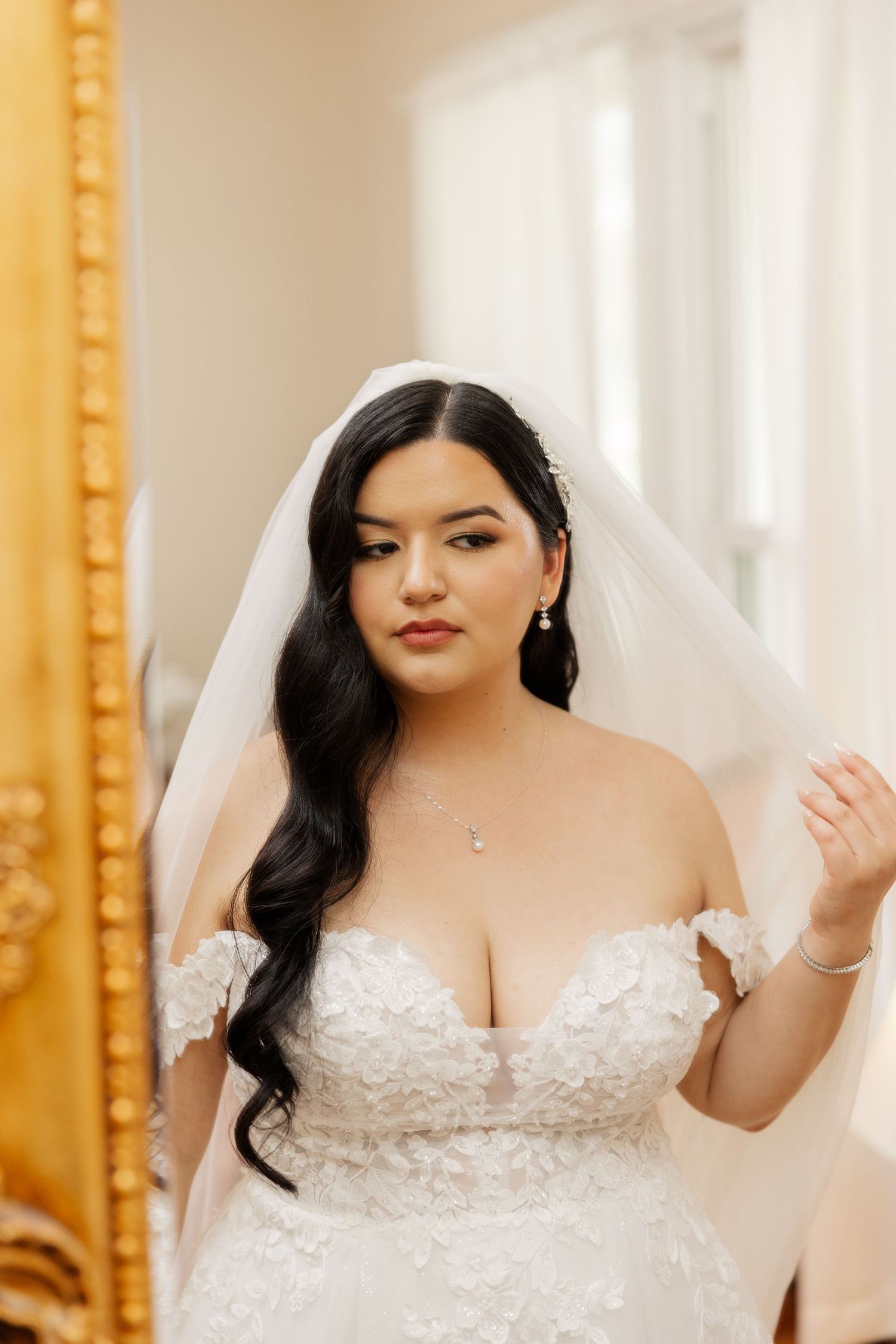 A bride wearing a beautiful off-shoulder lace wedding gown with a long veil and flowing dark hair poses elegantly.