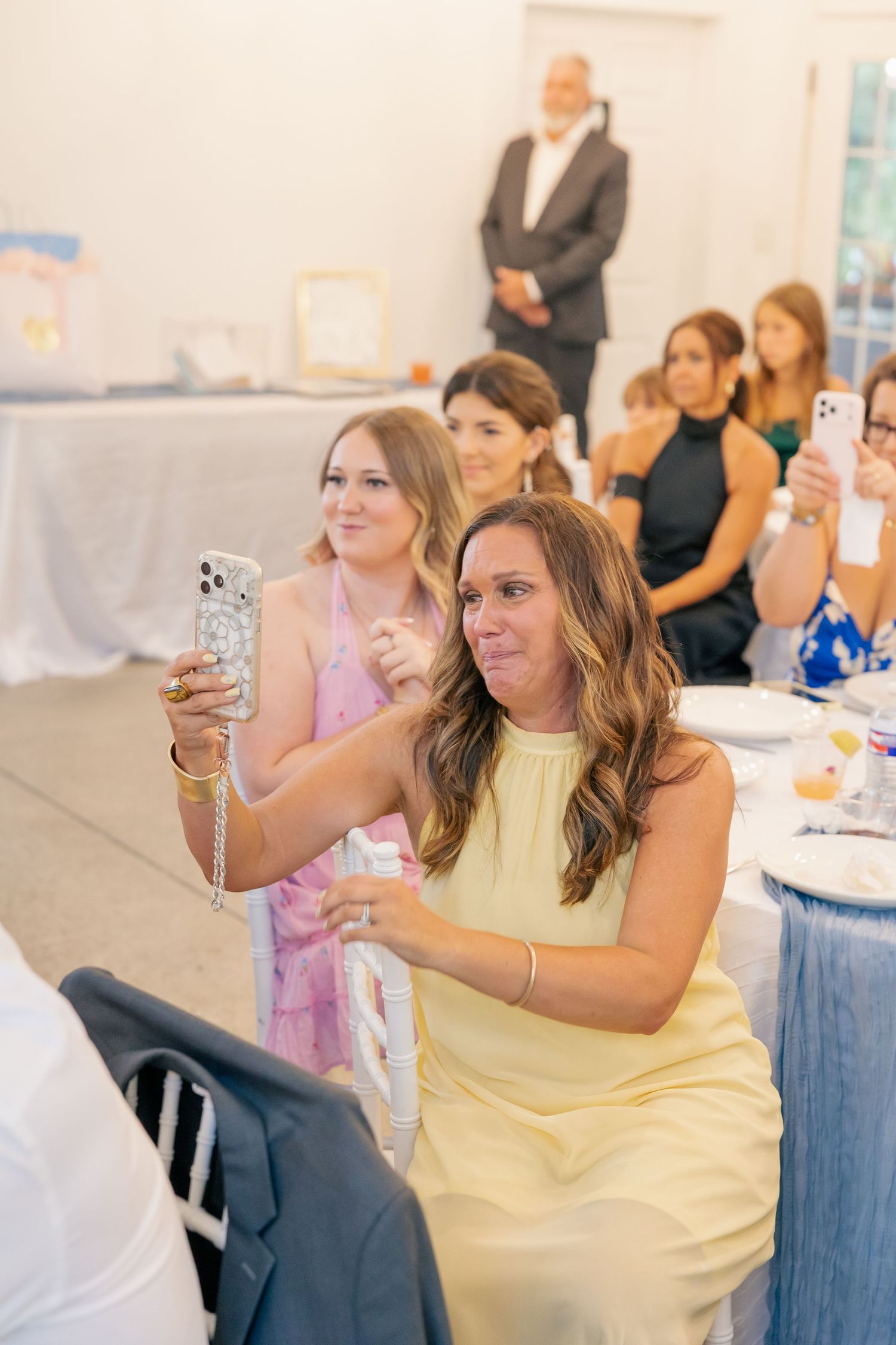 Wedding guests in colorful dresses photograph speeches on smartphones during elegant indoor reception.
