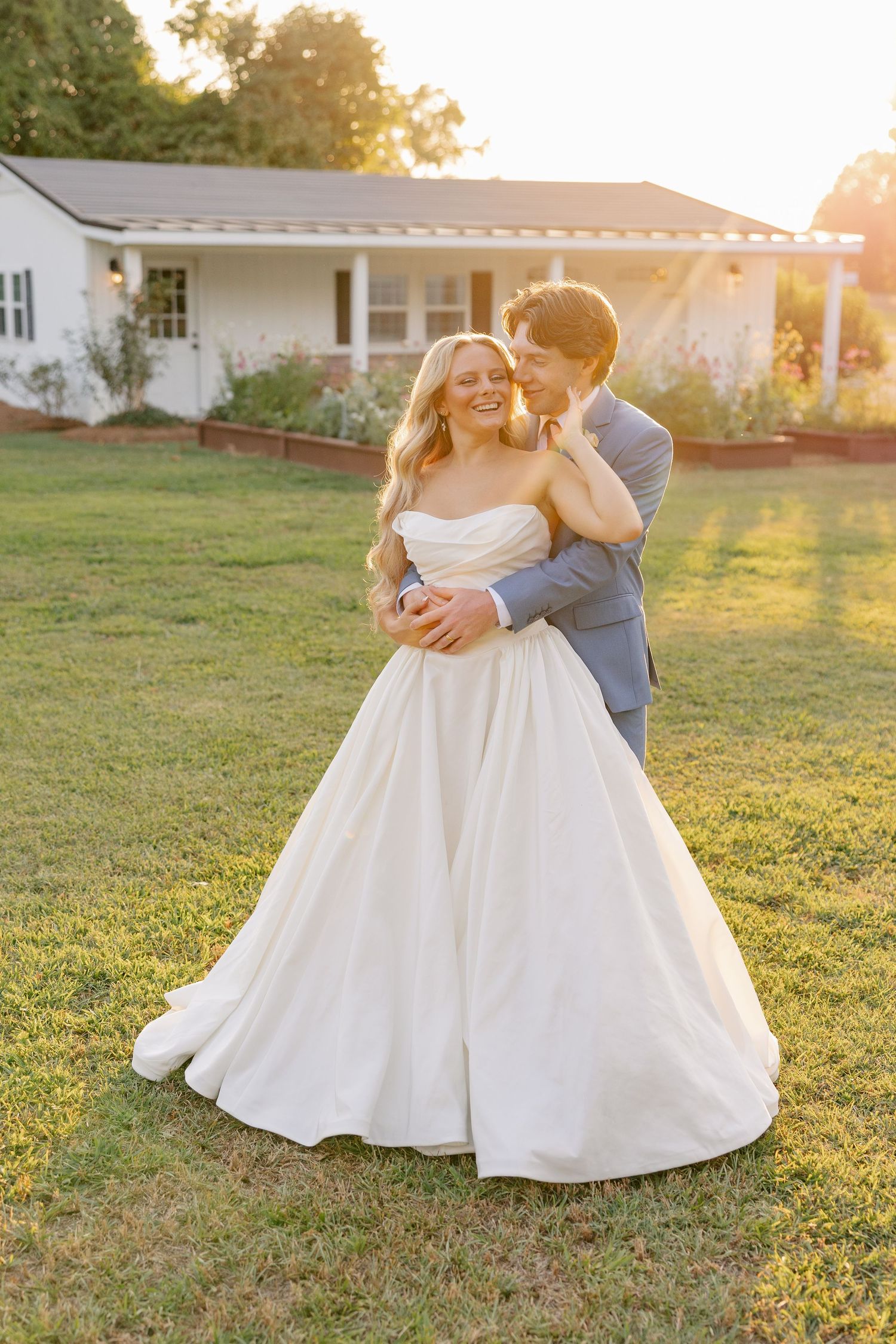 Groom in gray suit embraces smiling bride in strapless ballgown at sunset outside a white farmhouse venue.