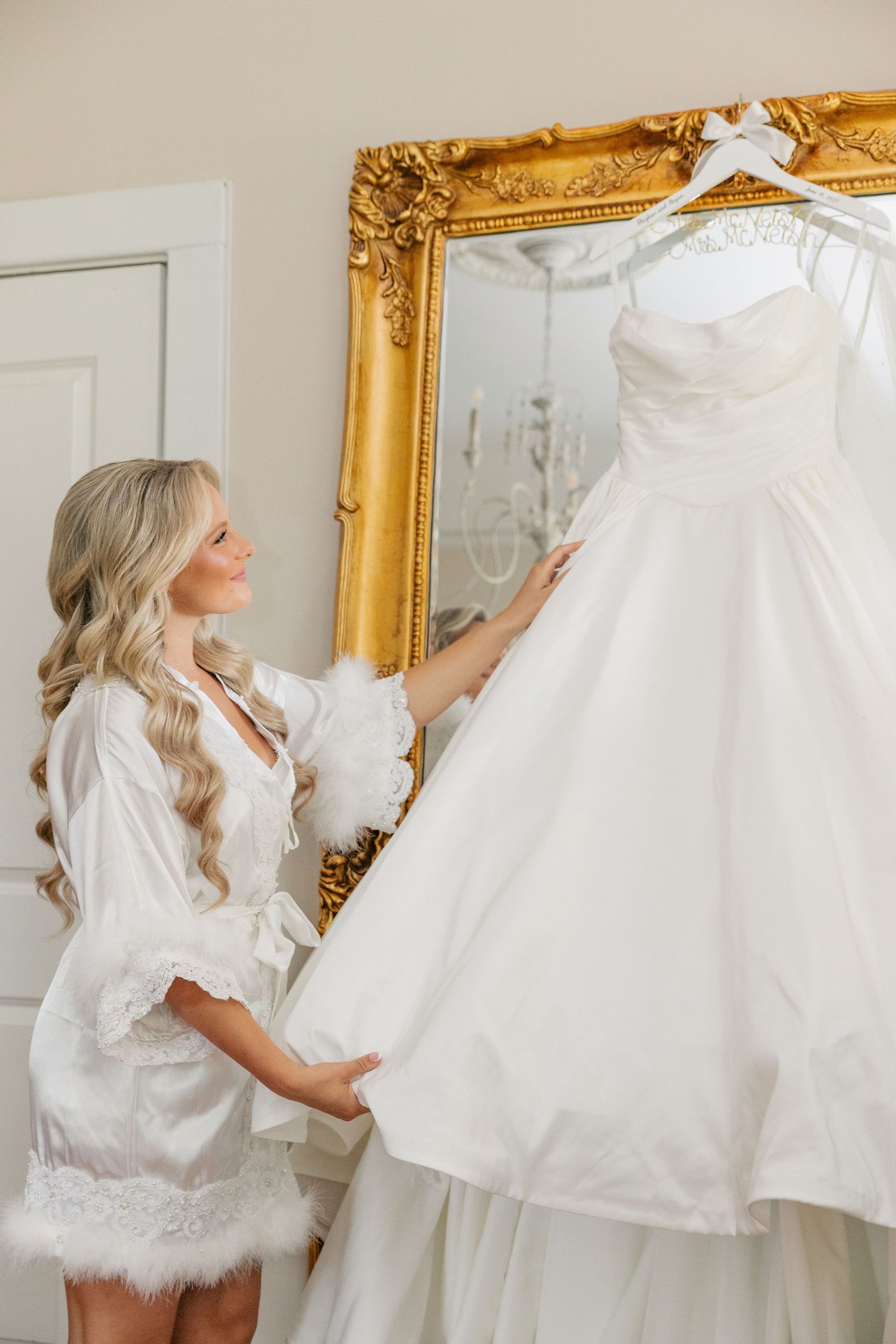 Bride in white lace robe admiring her wedding gown hanging on a gold ornate mirror.