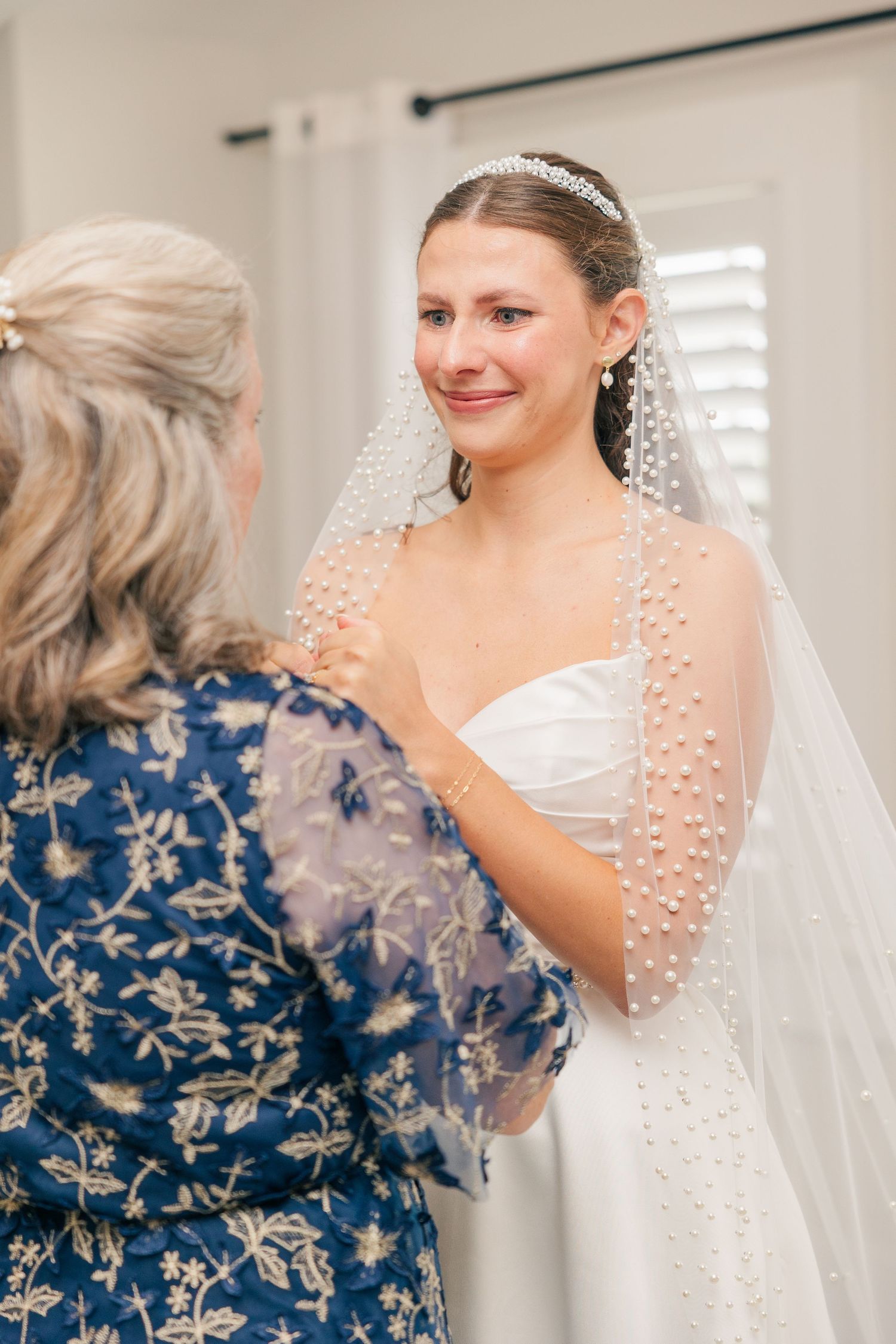 Bride smiling as older woman adjusts her pearl-adorned veil and white strapless wedding gown indoors.