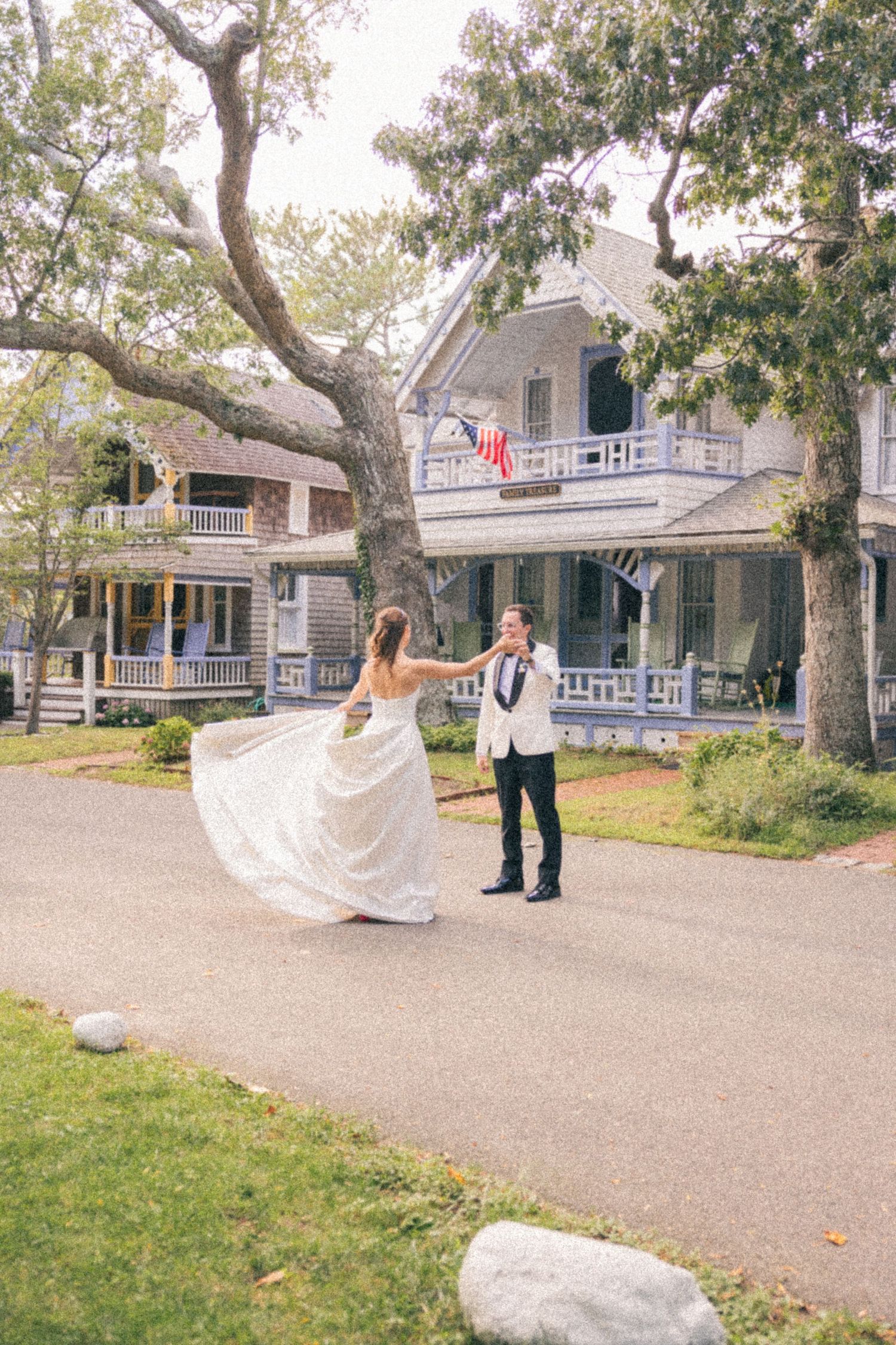 Bride in white gown twirls as groom watches on a tree-lined street in front of charming Victorian homes.
