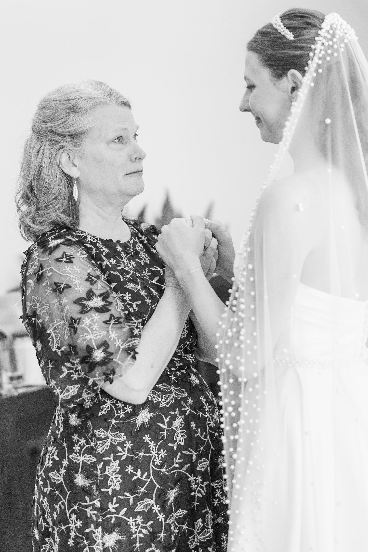 Black and white photo of mother holding hands with bride in pearl-trimmed veil on wedding day.