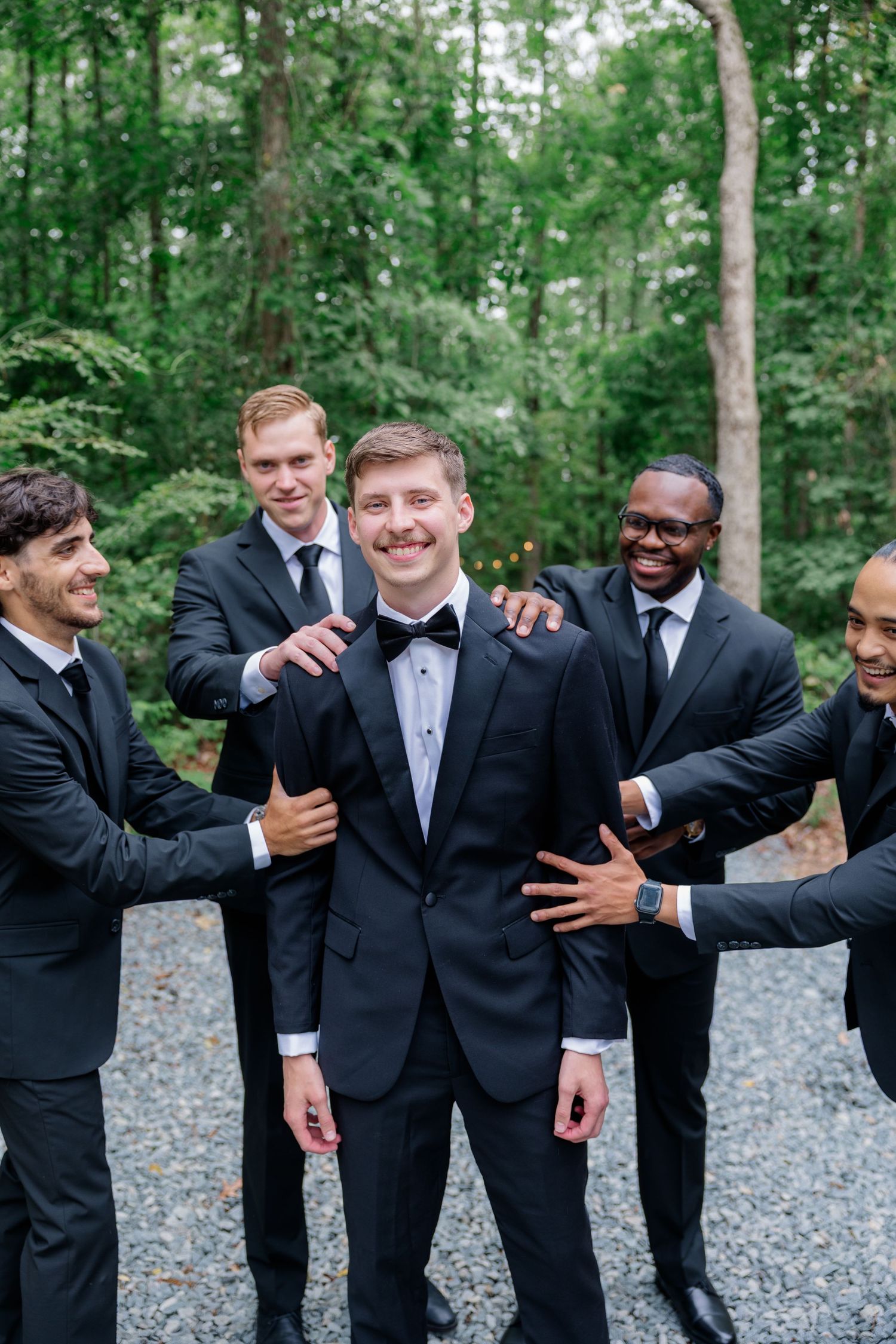 Groomsmen in black suits share laughs and embraces during candid moments at an outdoor wedding photoshoot.