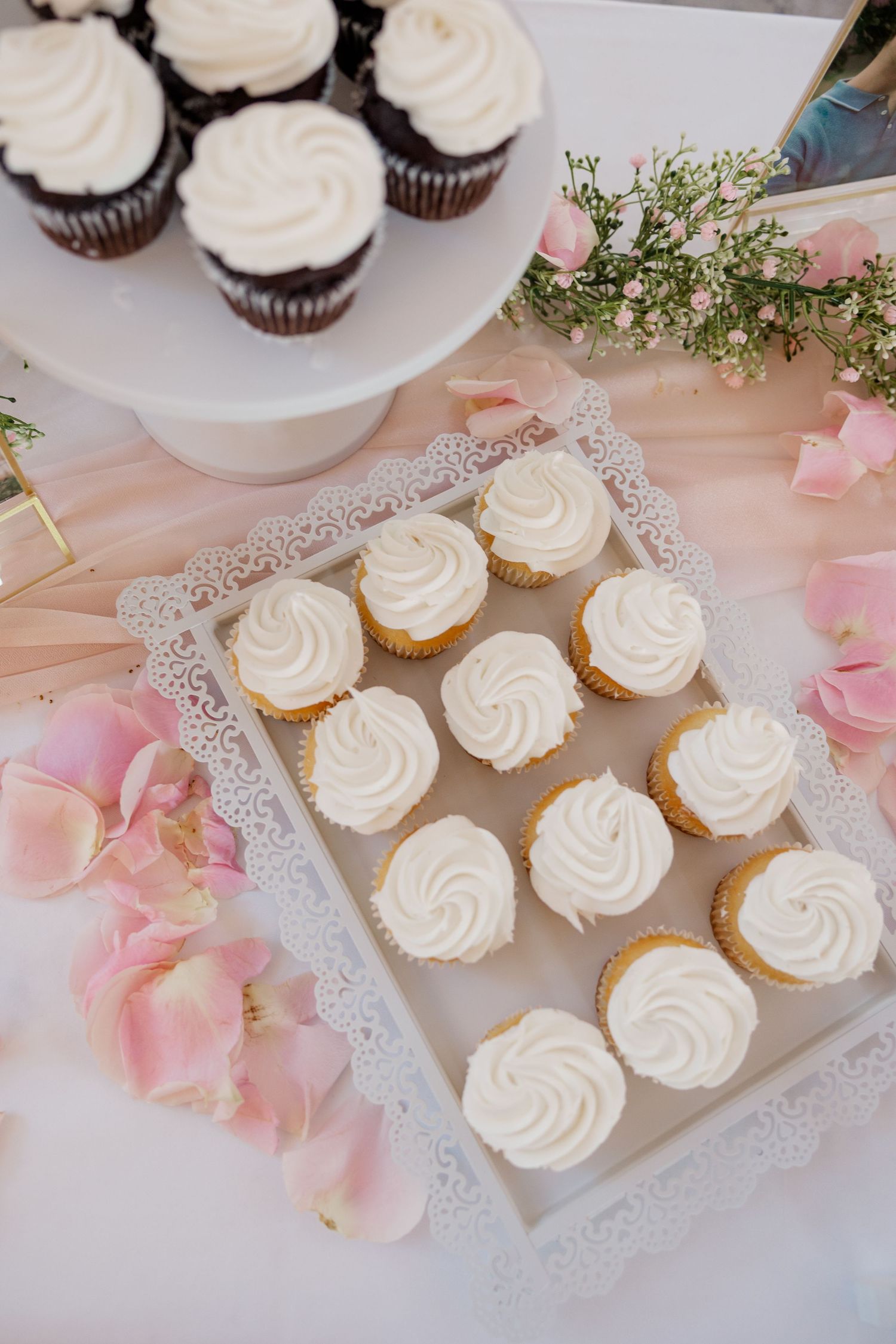 White frosted cupcakes on lace doilies decorated with pink flower petals for wedding reception.
