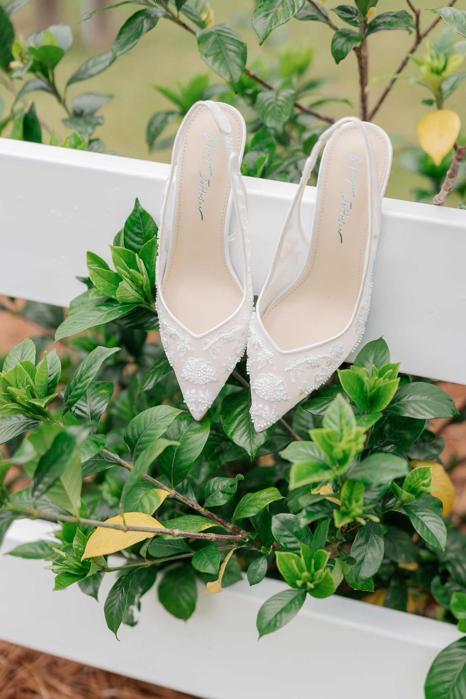 Elegant white lace bridal heels displayed on a white fence surrounded by lush green foliage.