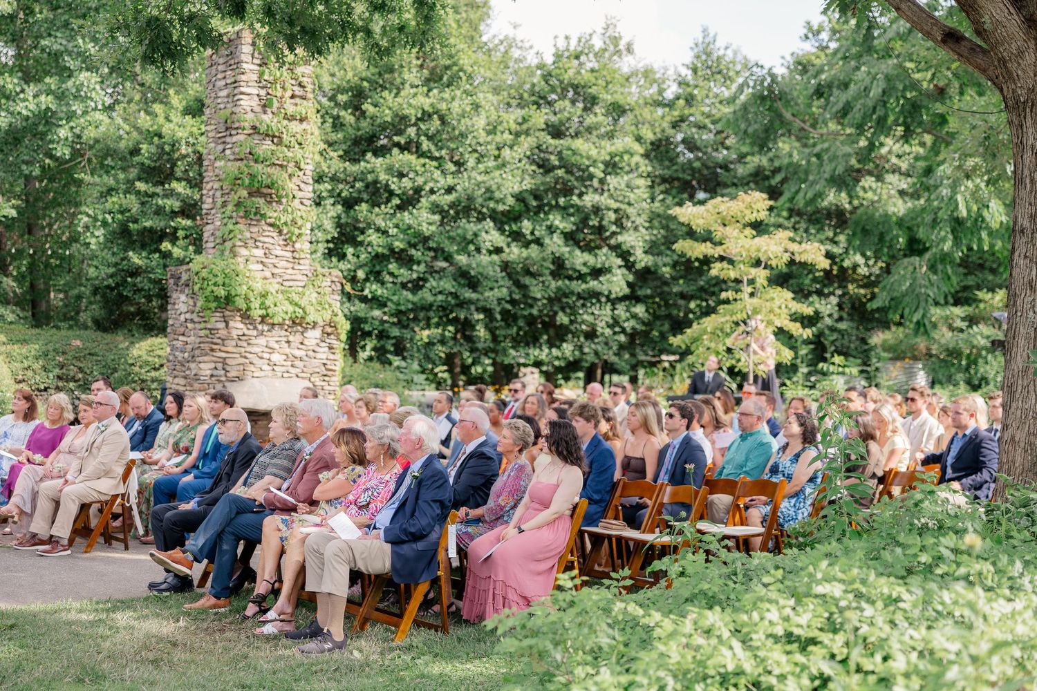 Outdoor garden wedding ceremony with guests seated in wooden chairs surrounded by lush greenery.