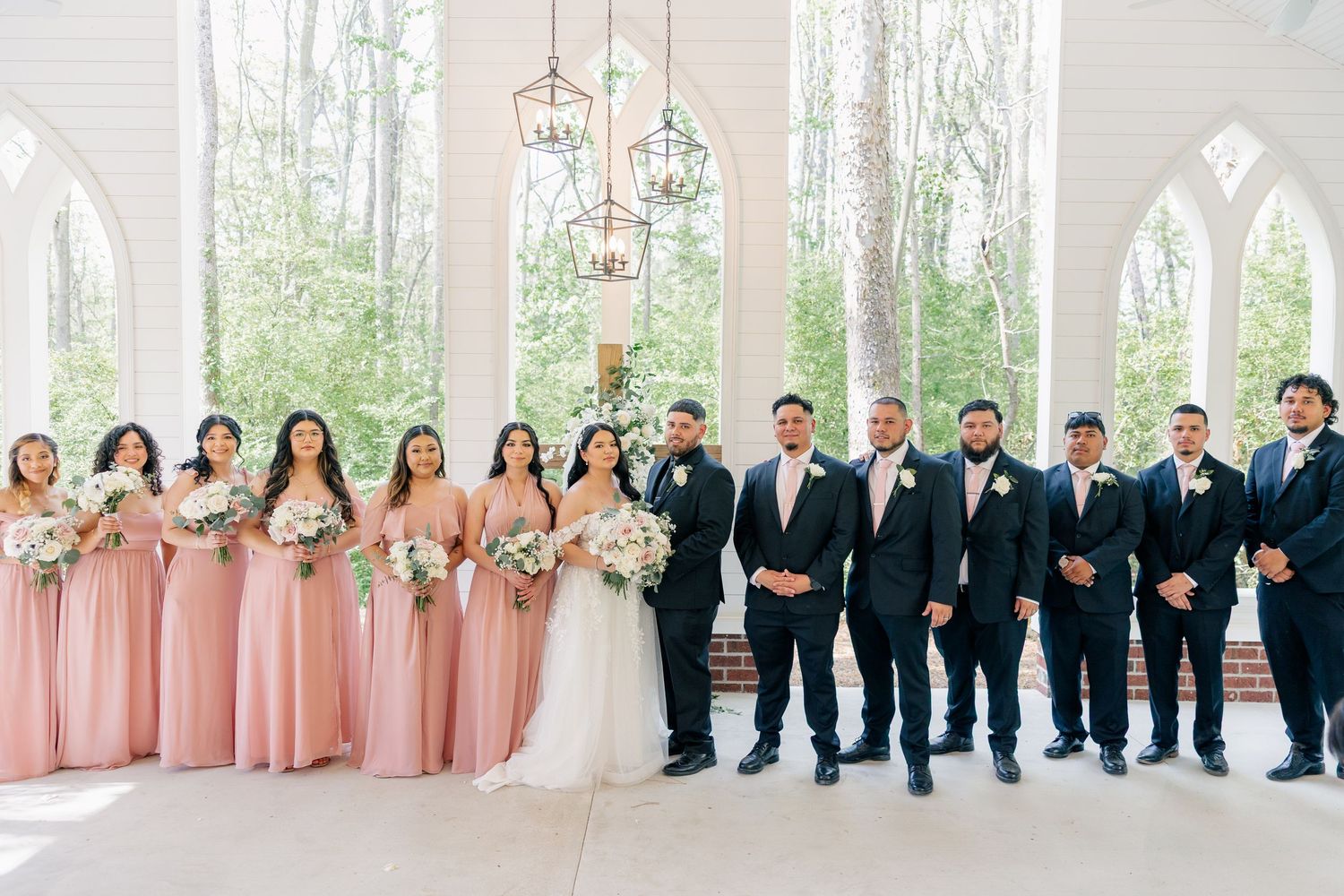 Wedding party members in pink dresses and black suits pose in a light-filled venue.