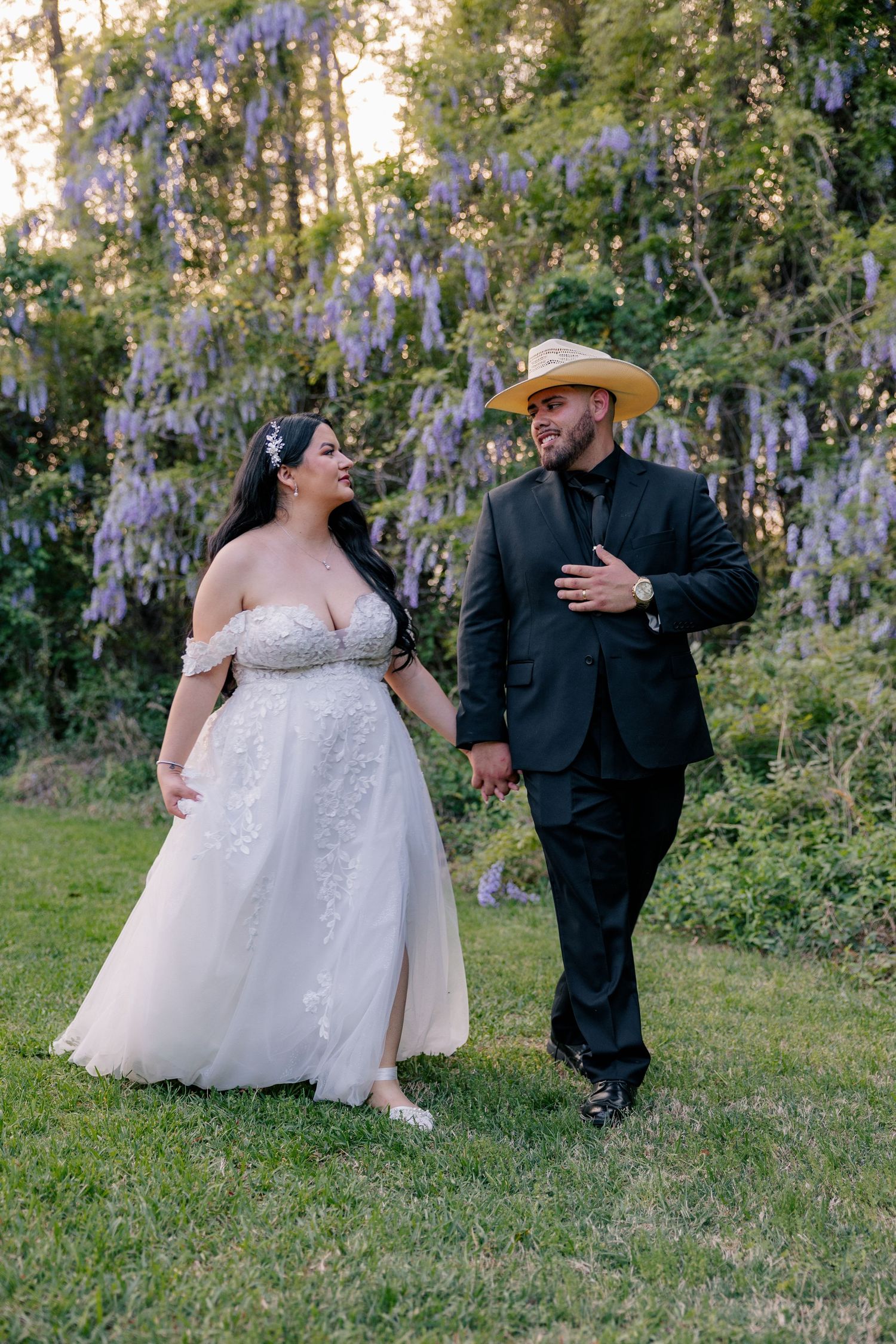 Couple in wedding attire share candid moments while sitting in a garden with blooming purple wisteria.