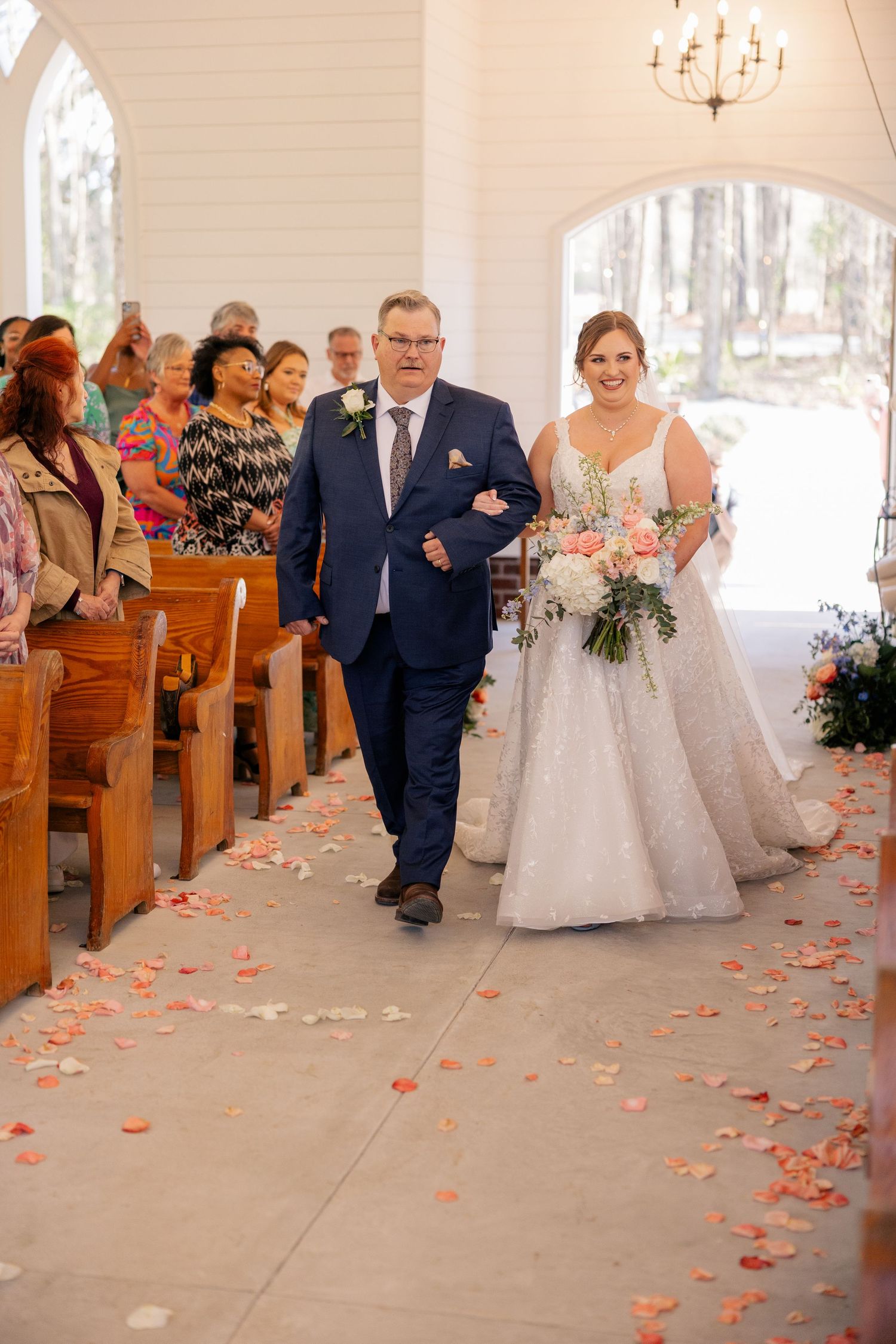 Wedding ceremony in progress inside a bright chapel with coral rose petals lining the aisle.
