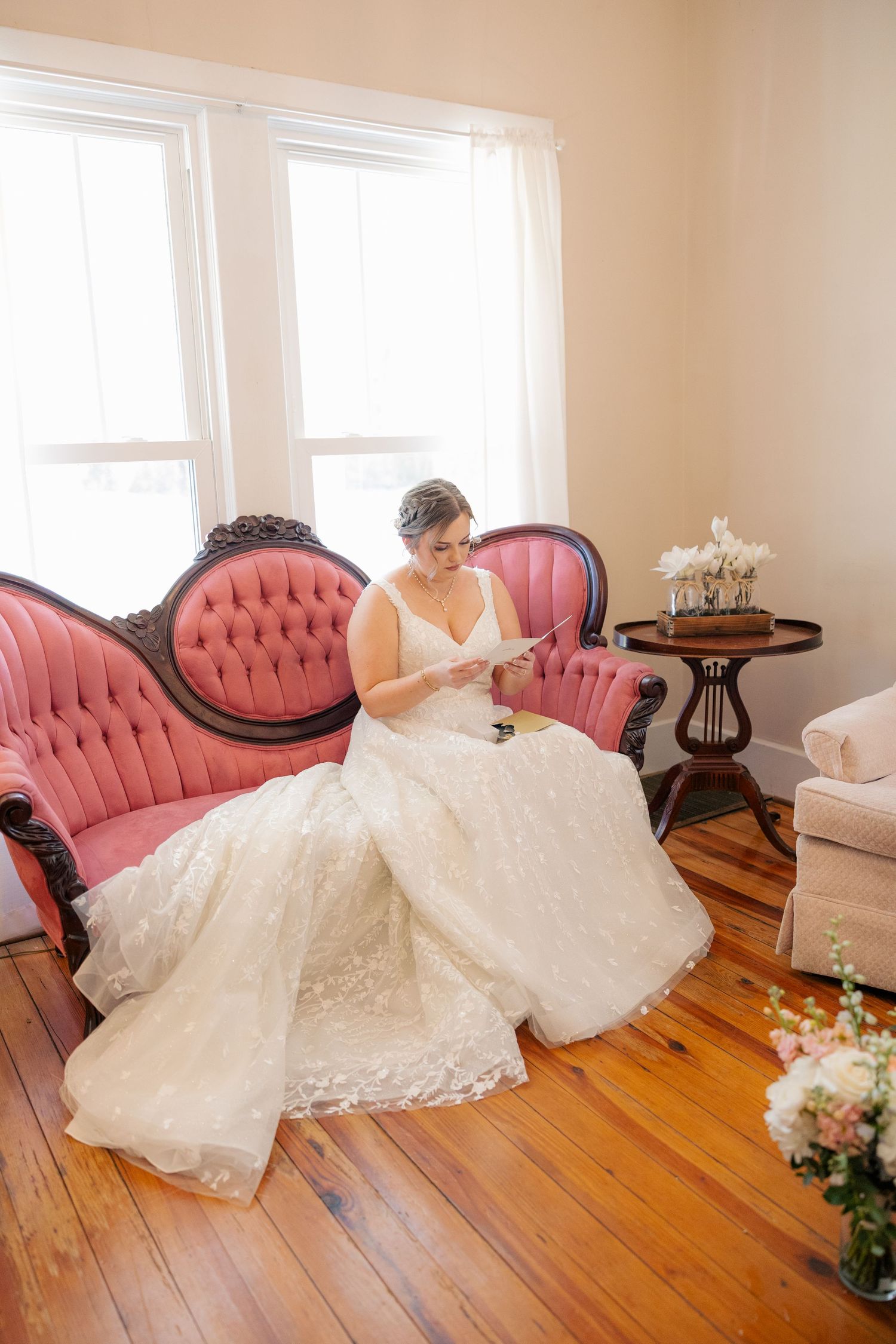 A white wedding dress with lace details spreads elegantly across a hardwood floor with pink antique chairs in the background.