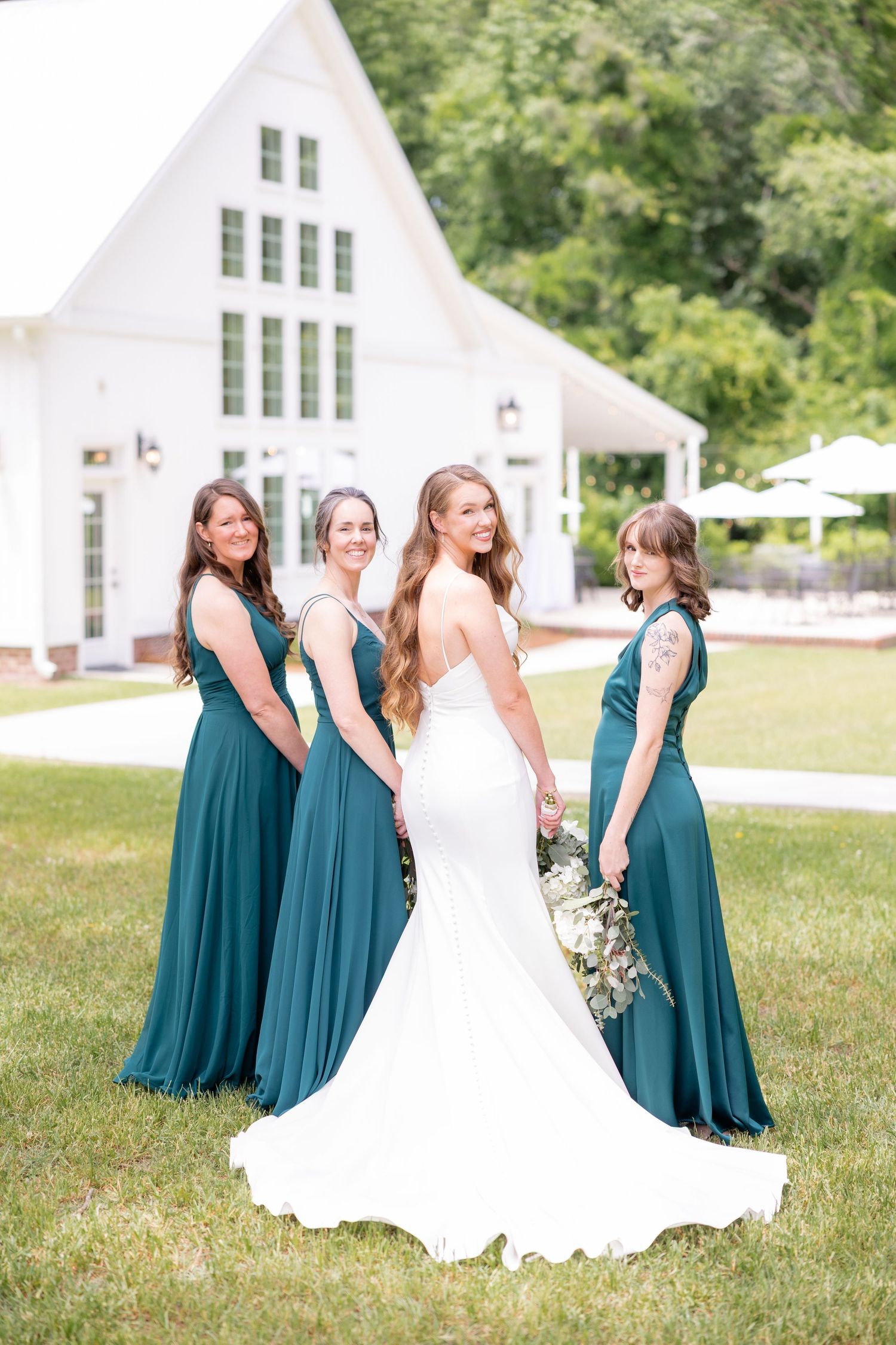Wedding party in teal bridesmaid dresses and white gown pose on grassy lawn in front of white chapel.