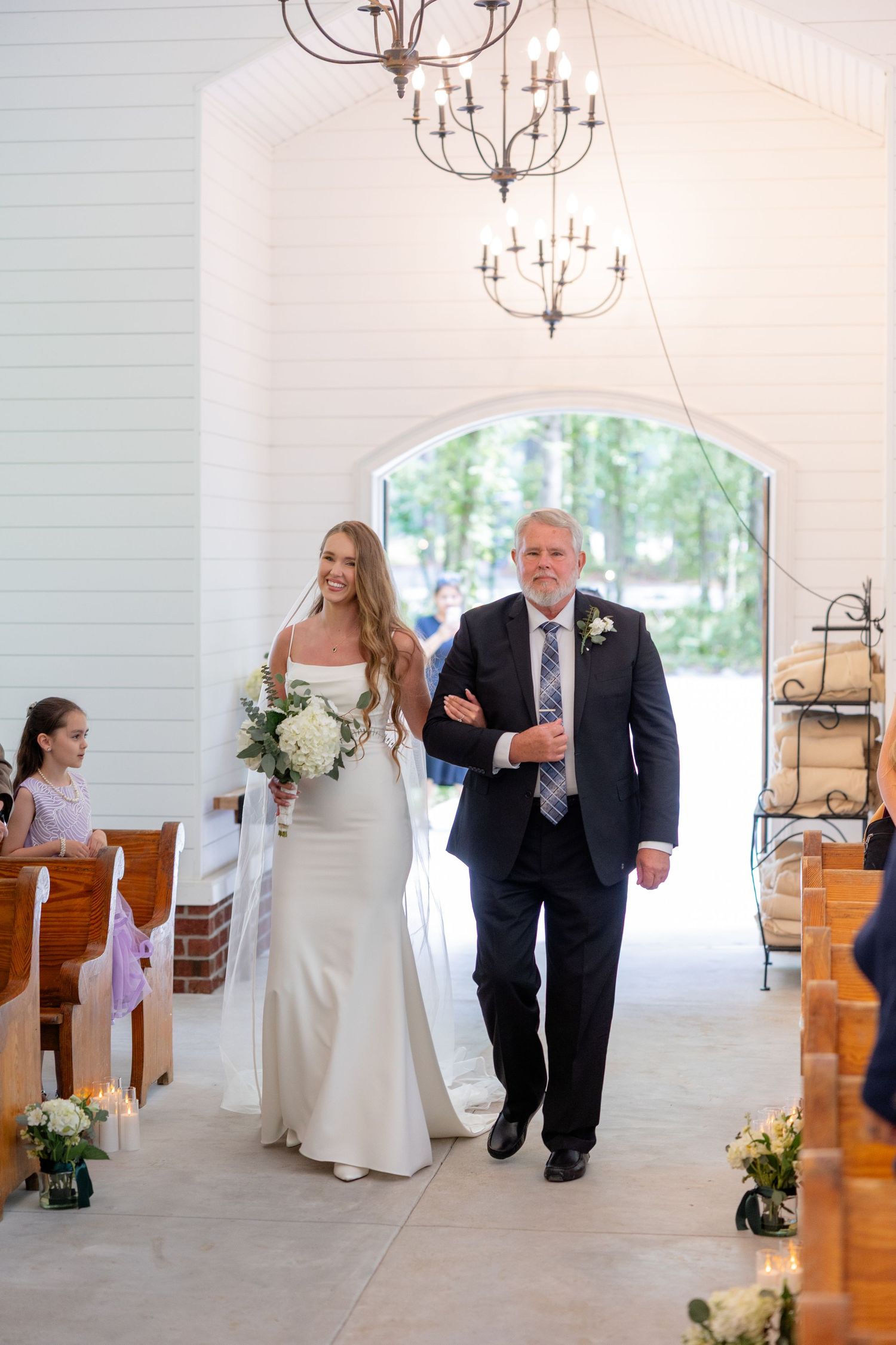 A bride and her escort walking through an elegant white chapel with chandeliers overhead.