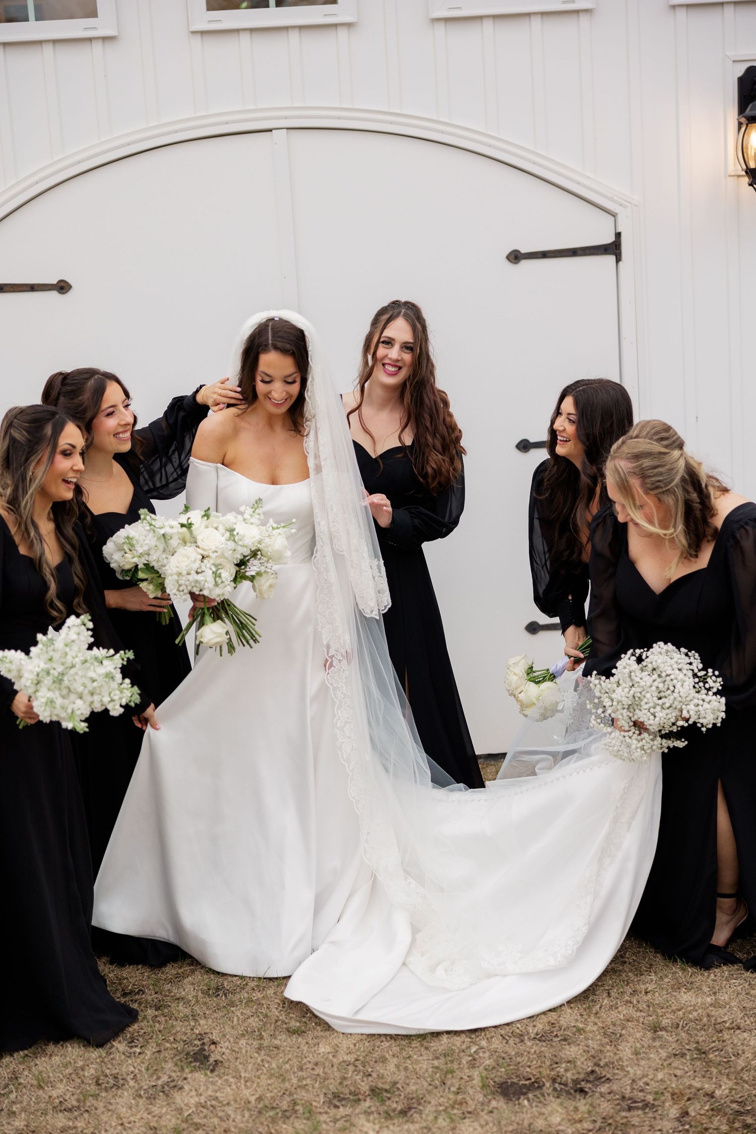 Bridesmaids in black dresses pose playfully with a bride in white against a white barn wall.