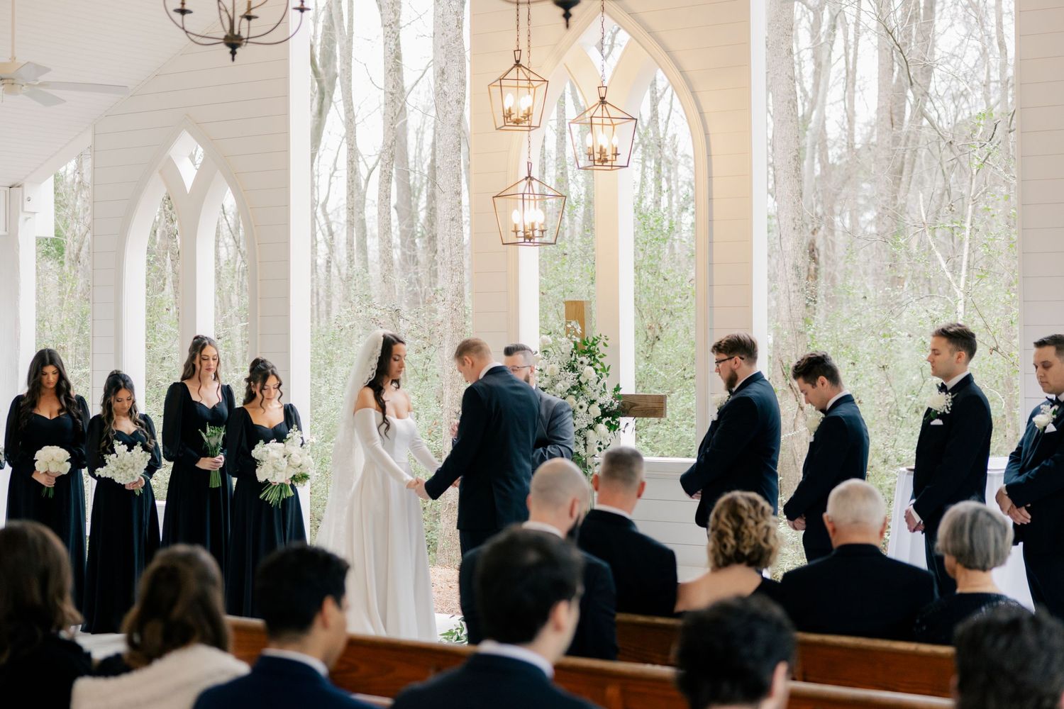 Wedding guests gather inside a bright white chapel with large windows and elegant chandeliers for a ceremony.