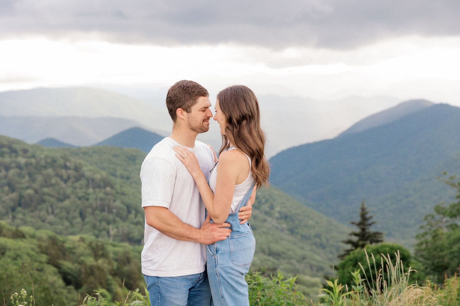 A couple embraces while standing on a mountain overlook with a scenic valley and green hills in the background.