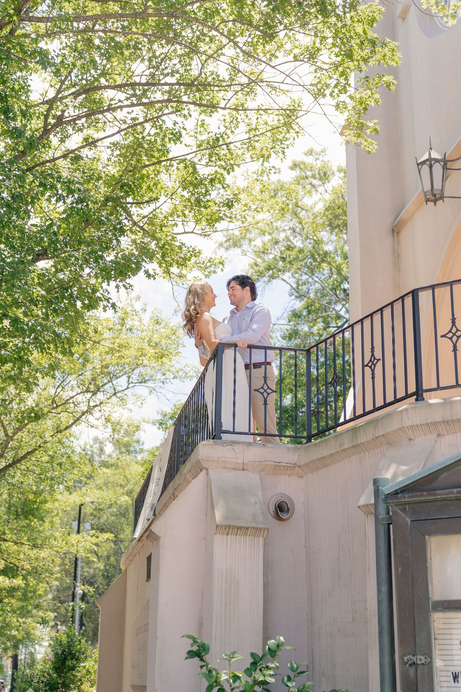 Couple standing on a balcony of a beige building surrounded by lush green trees on a sunny day.