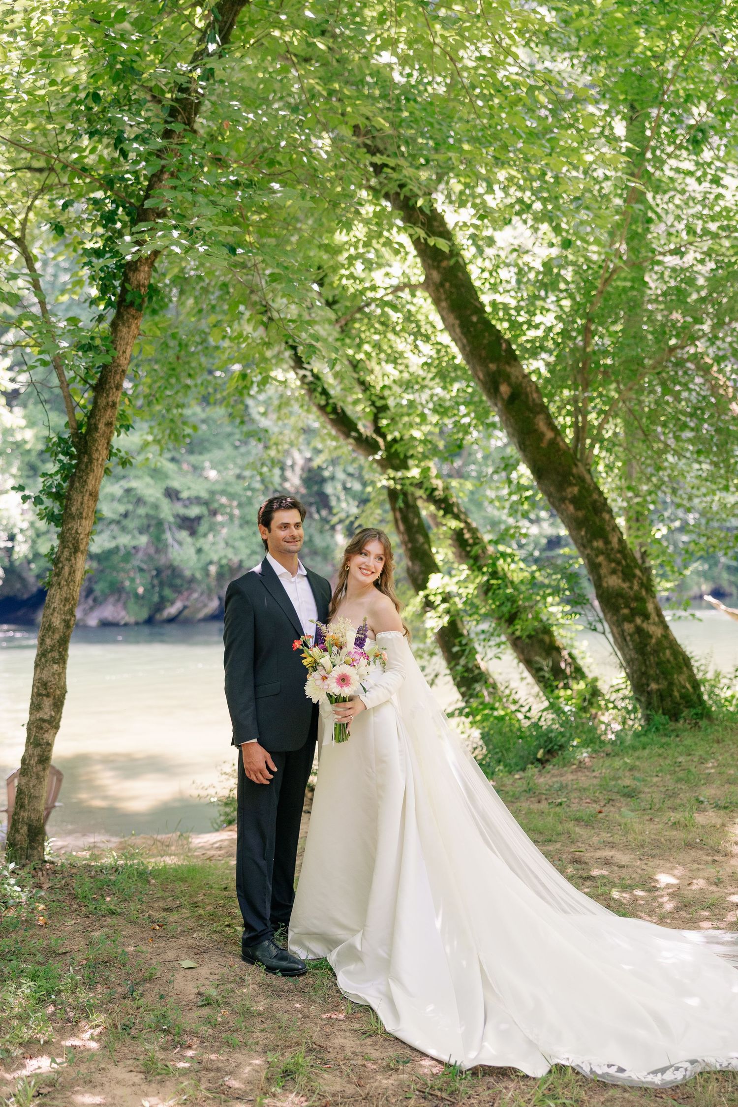 Bride and groom standing under lush green trees by a river, bride holding a colorful wedding bouquet.