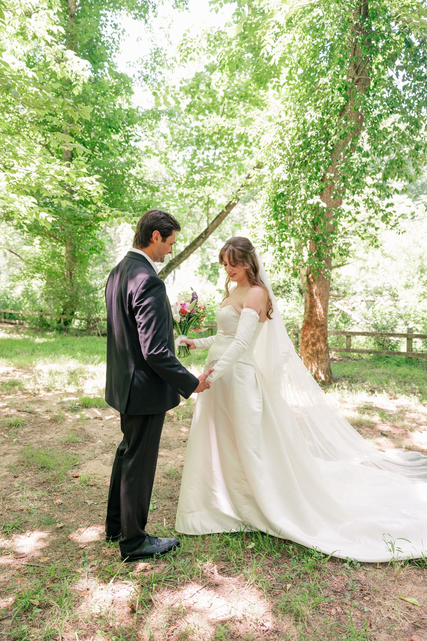 Bride and groom share a tender first look moment holding hands in a sunlit forest setting.
