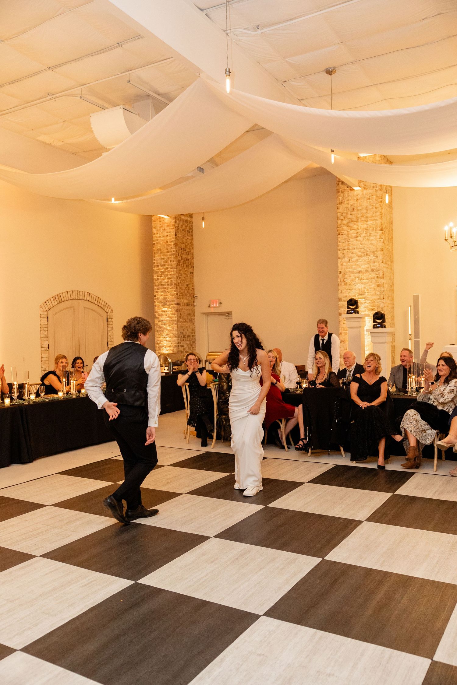 A wedding couple shares their first dance on a black and white checkered floor under white fabric ceiling drapes.
