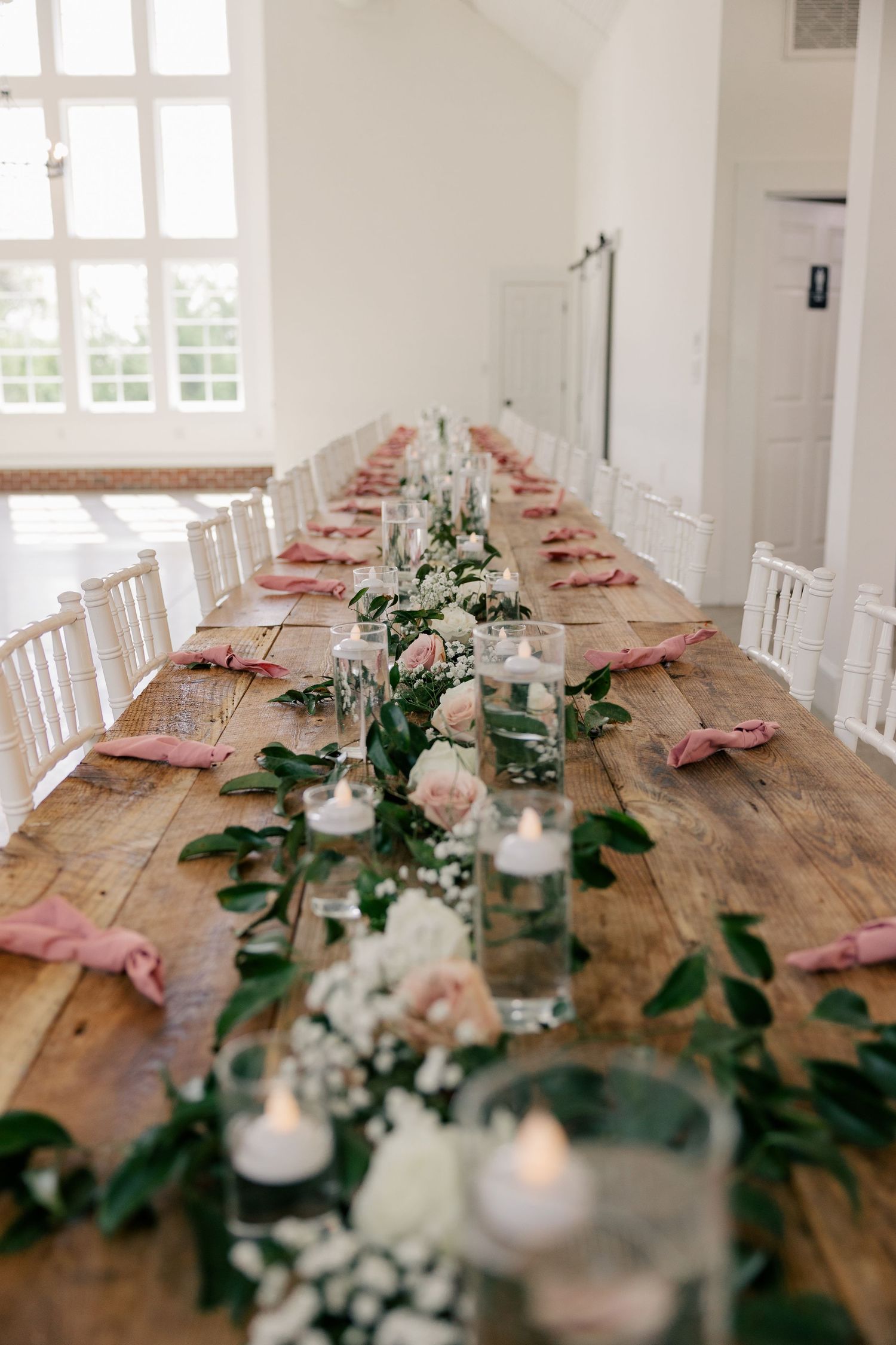 An elegant rustic wedding reception table with white chairs, blush florals, and candlelight on a wooden farmhouse table.
