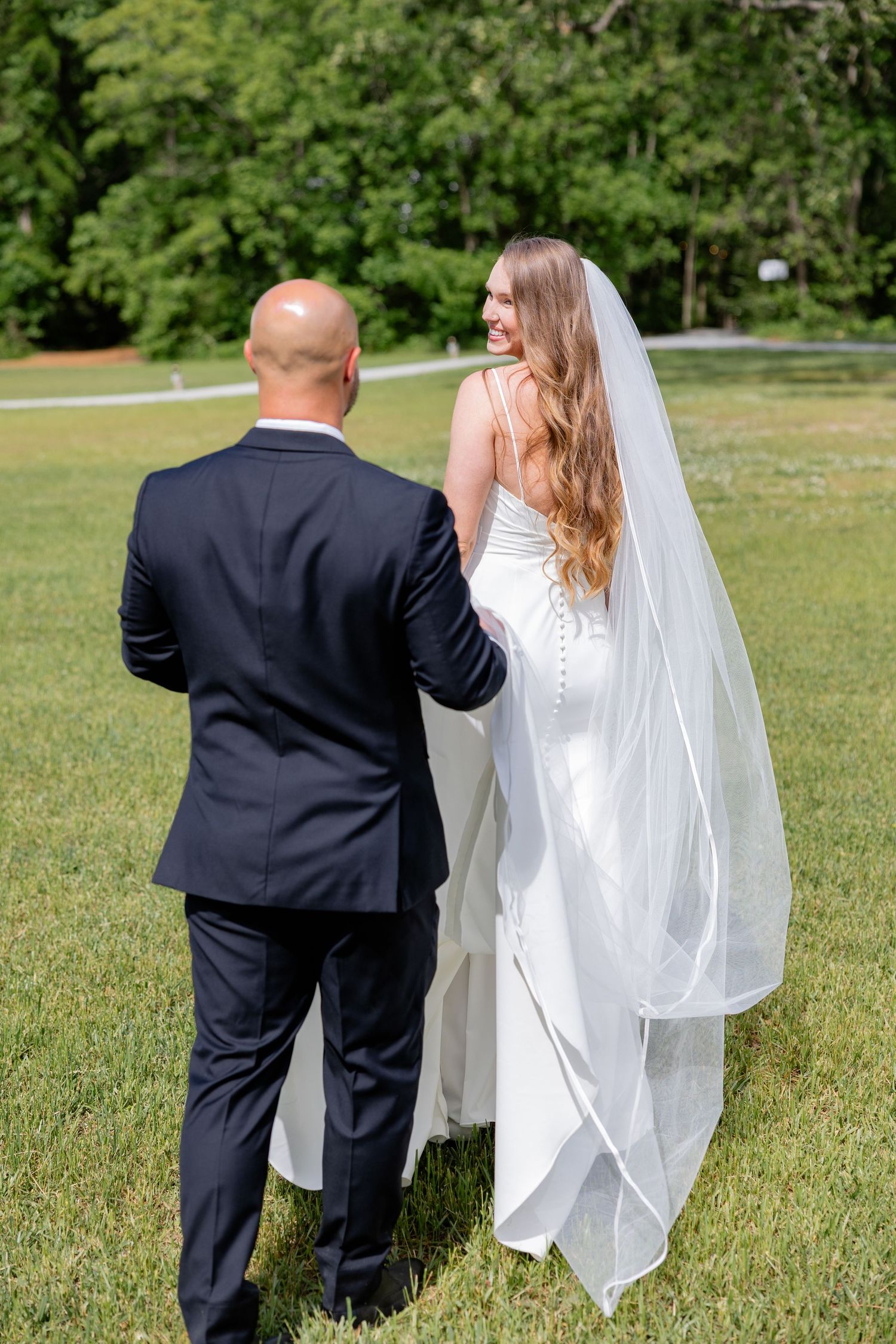 A couple in wedding attire stand together on a grassy lawn with the bride wearing a flowing veil.