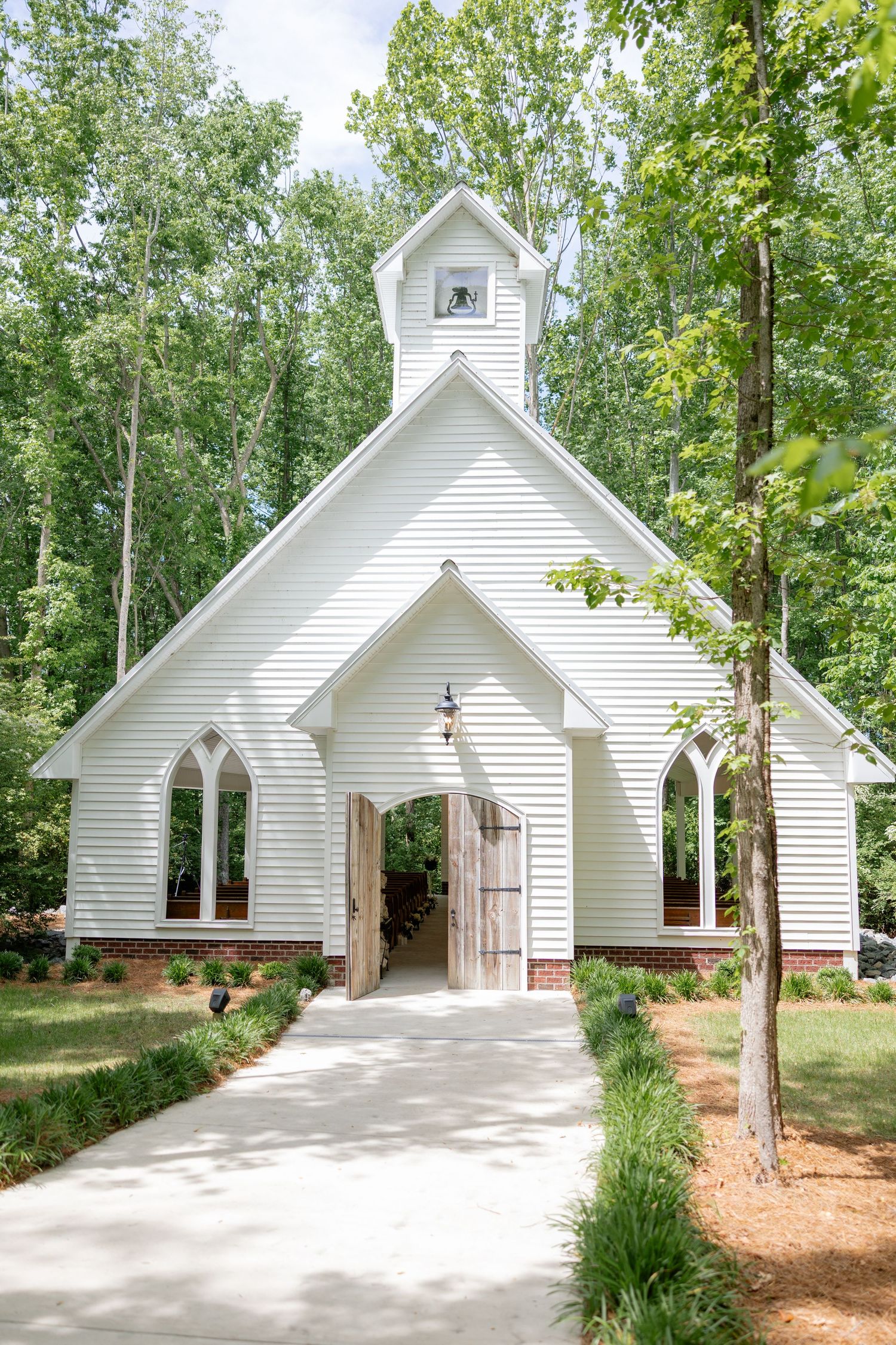 A white chapel with gothic arched windows and a concrete walkway sits nestled among lush green trees.