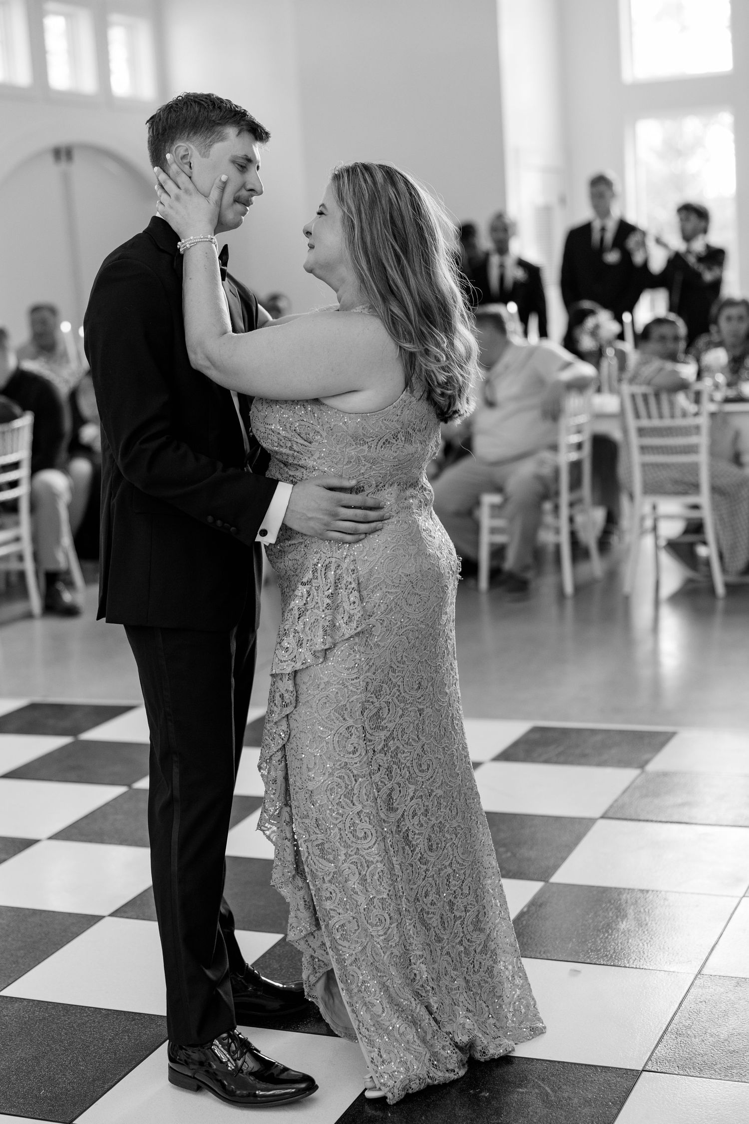 A couple dancing together at a formal event on a checkered floor, the woman wearing a sparkly long dress.