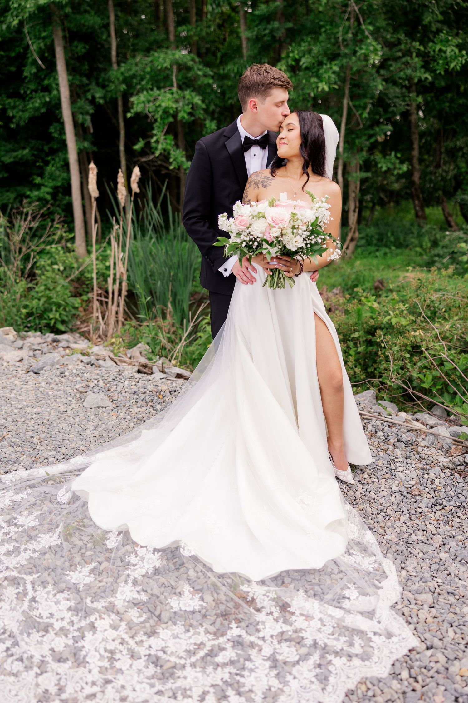Elegant wedding couple poses outdoors holding bouquet of pink roses against natural backdrop.