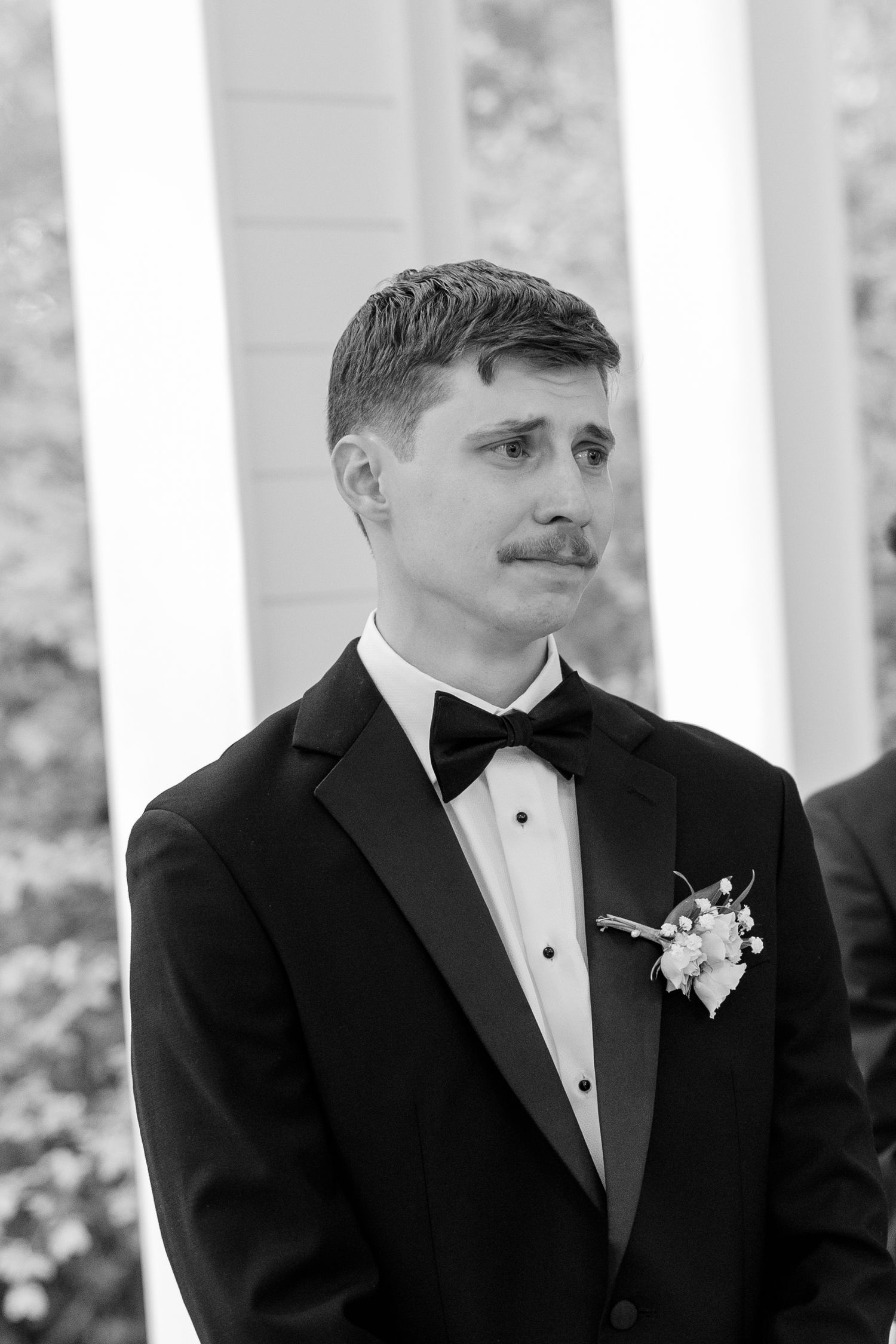 Close-up shots of a groom in a black tuxedo and white boutonniere standing in front of white pillars.