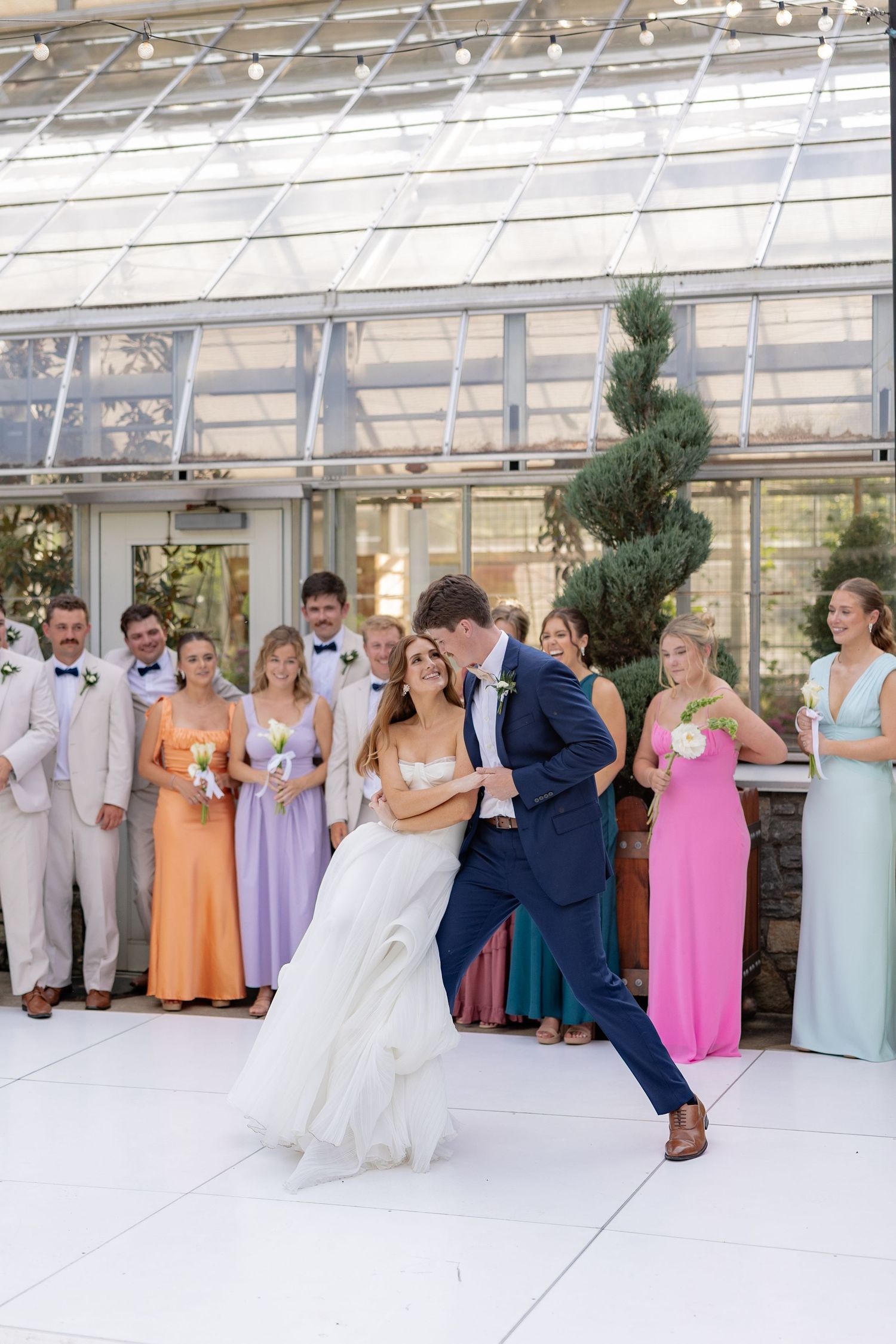 Wedding party lined up in a greenhouse with bridesmaids in pastel dresses and groomsmen in light suits.