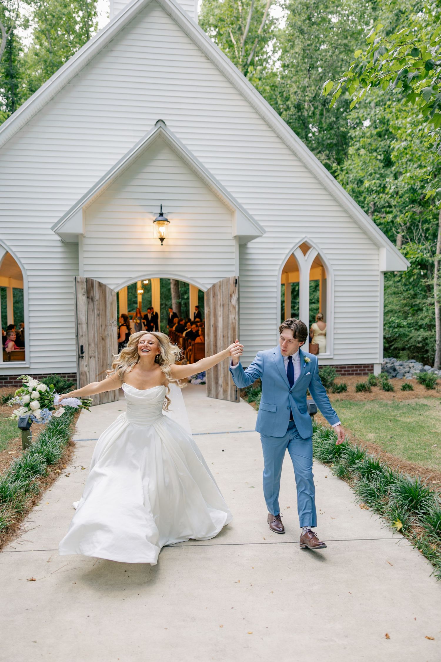 Bride and groom celebrating joyfully outside white chapel after wedding ceremony at wooded venue.