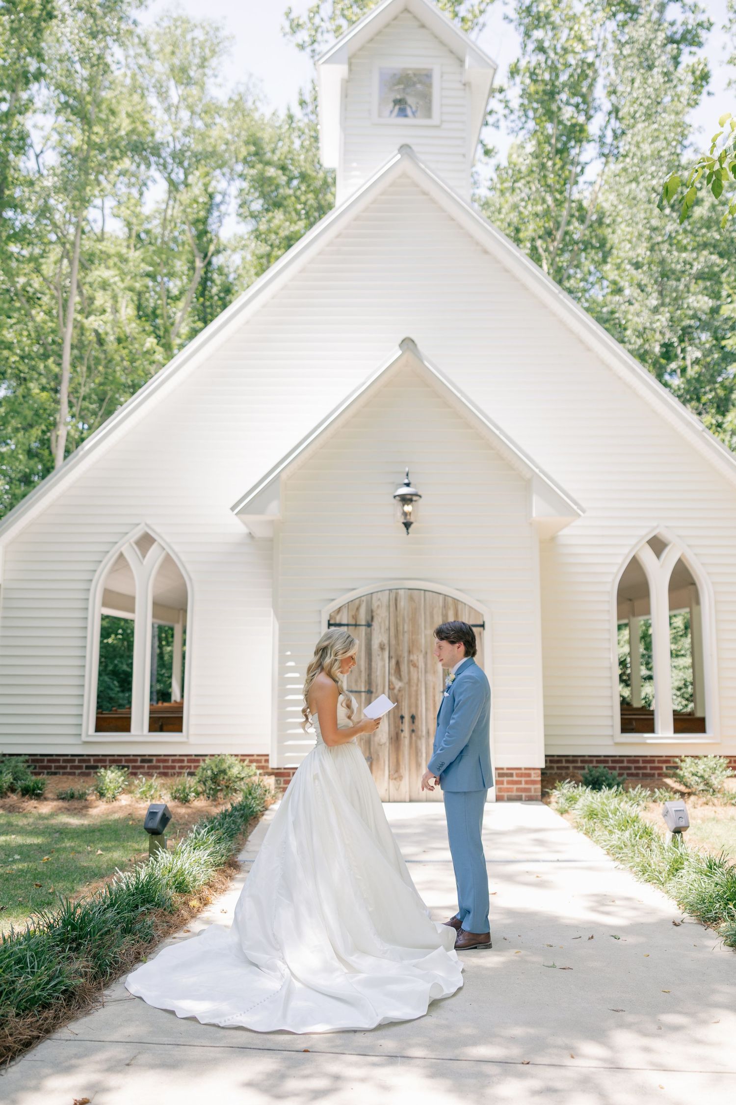 Bride and groom exchange vows outside a charming white chapel surrounded by lush green trees.