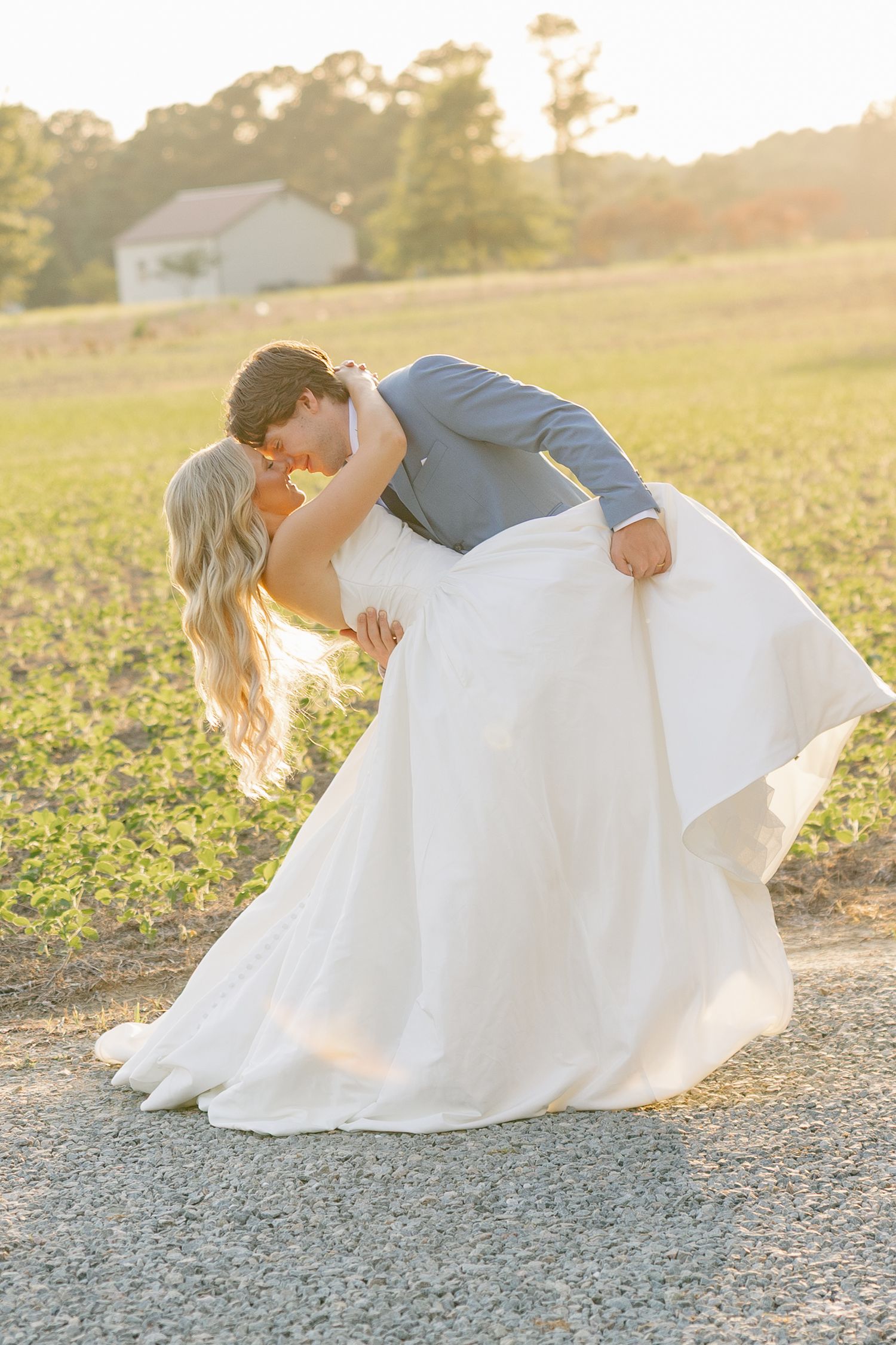 Groom dips bride in romantic golden hour kiss on gravel path beside a sunlit farm field.