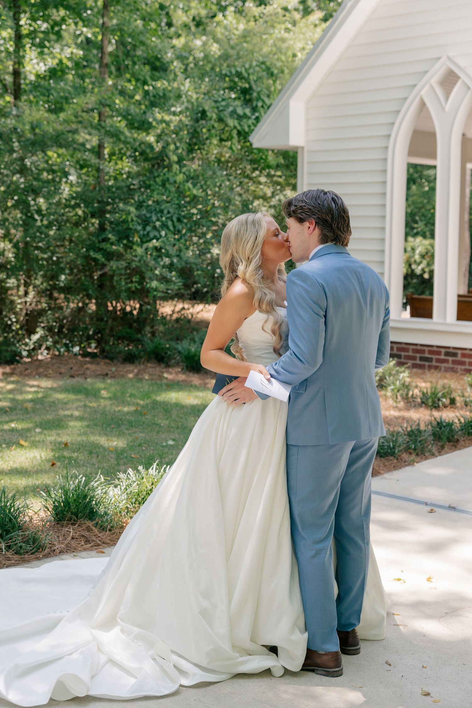 Bride and groom share a romantic kiss outside a white chapel surrounded by lush green trees.