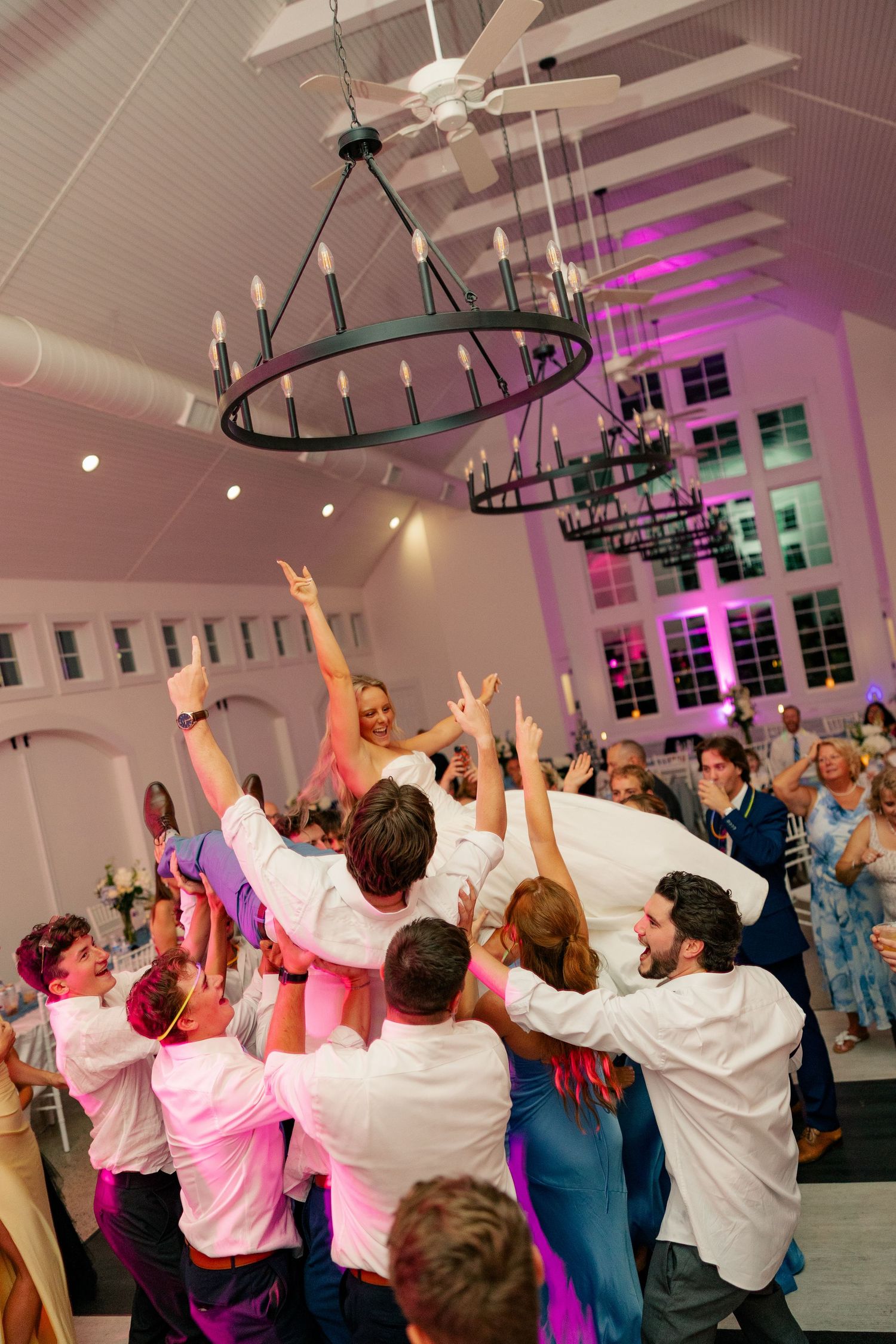 Guests lift bride at lively wedding reception under chandelier lights.