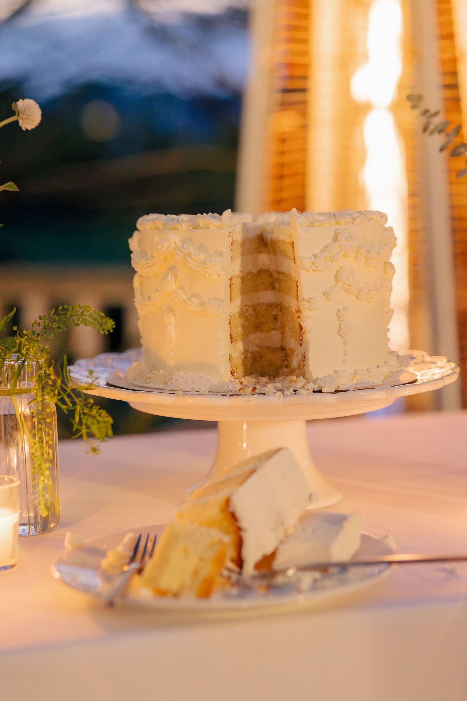 Elegant white layered wedding cake on pedestal stand with slice served on plate.