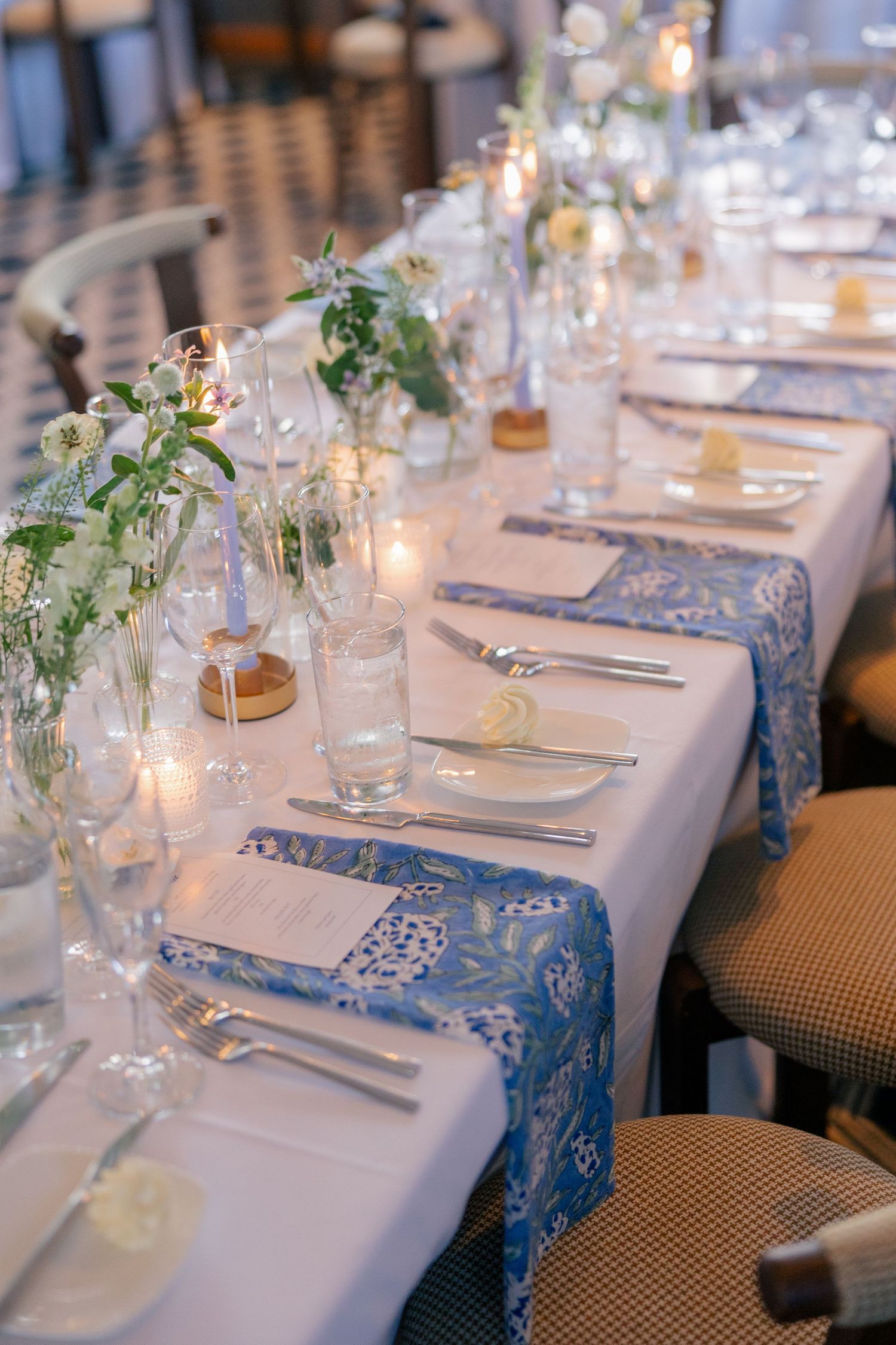 Elegant wedding reception table with blue floral runners, white linens, candles, and floral centerpieces.