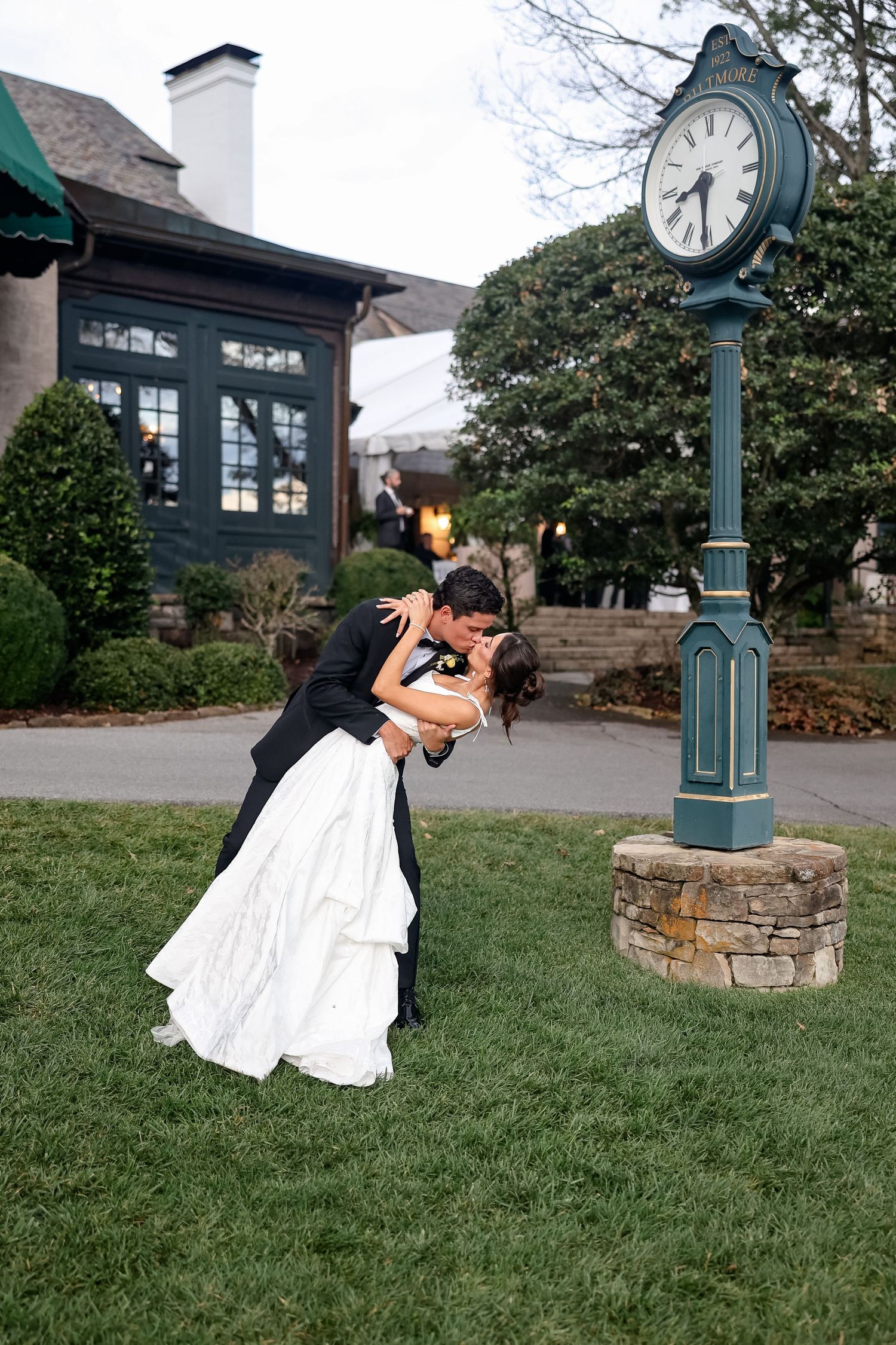 Bride and groom share a romantic dip kiss on green lawn beside vintage clock tower at elegant wedding venue.