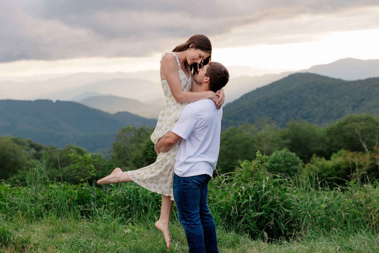 Couple shares an intimate moment in a grassy field with mountain views and dramatic clouds.